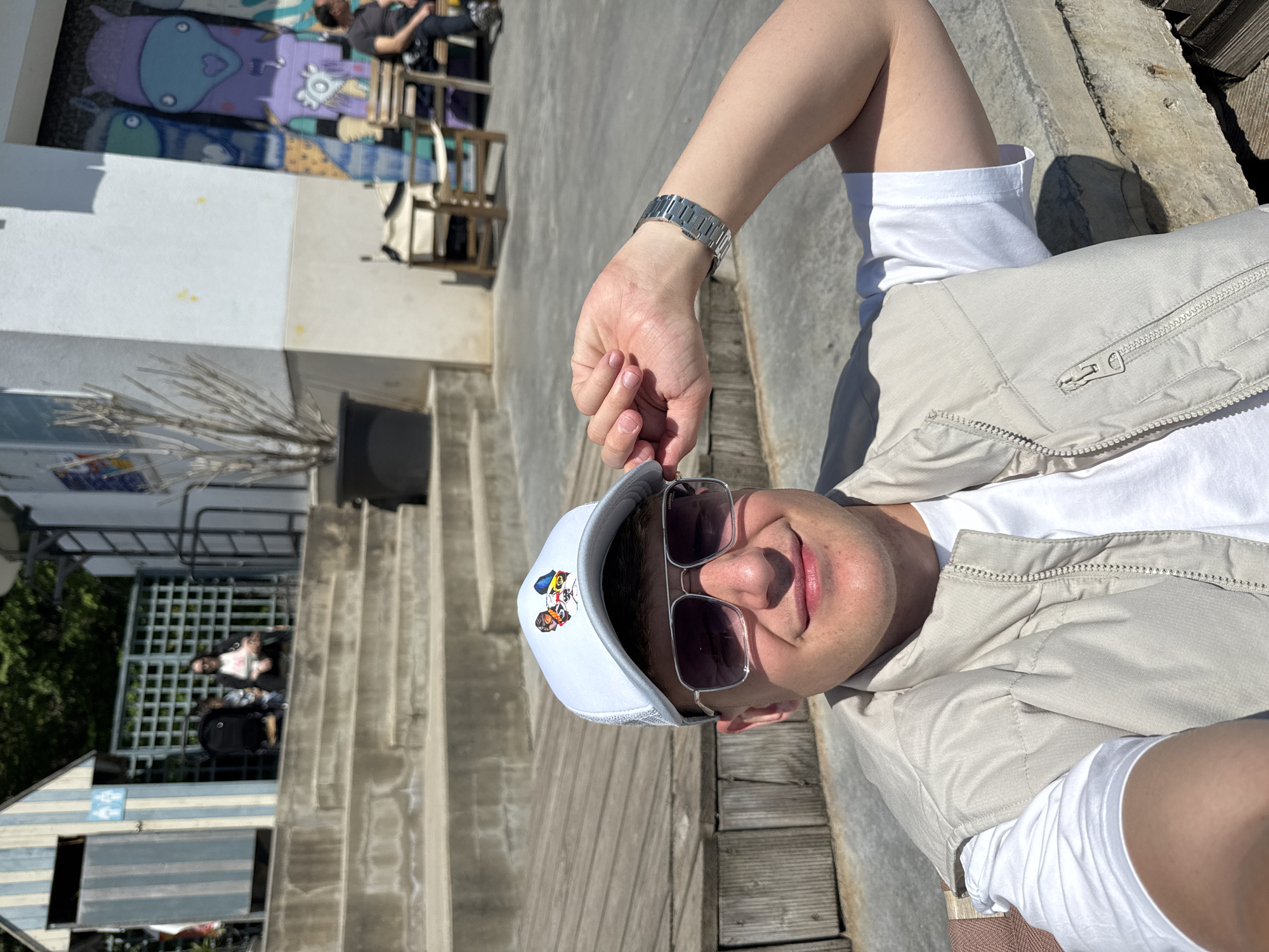 A man is posing for a selfie outside. The man is wearing a white t-shirt, a light-colored vest, a white cap with a cartoon design, and sunglasses. He is also wearing a silver watch on his left wrist. The background shows concrete steps, wooden planks, and a building with colorful graffiti art. A few people are visible sitting on chairs in the background. The location appears to be an outdoor public space in Varna, Bulgaria.