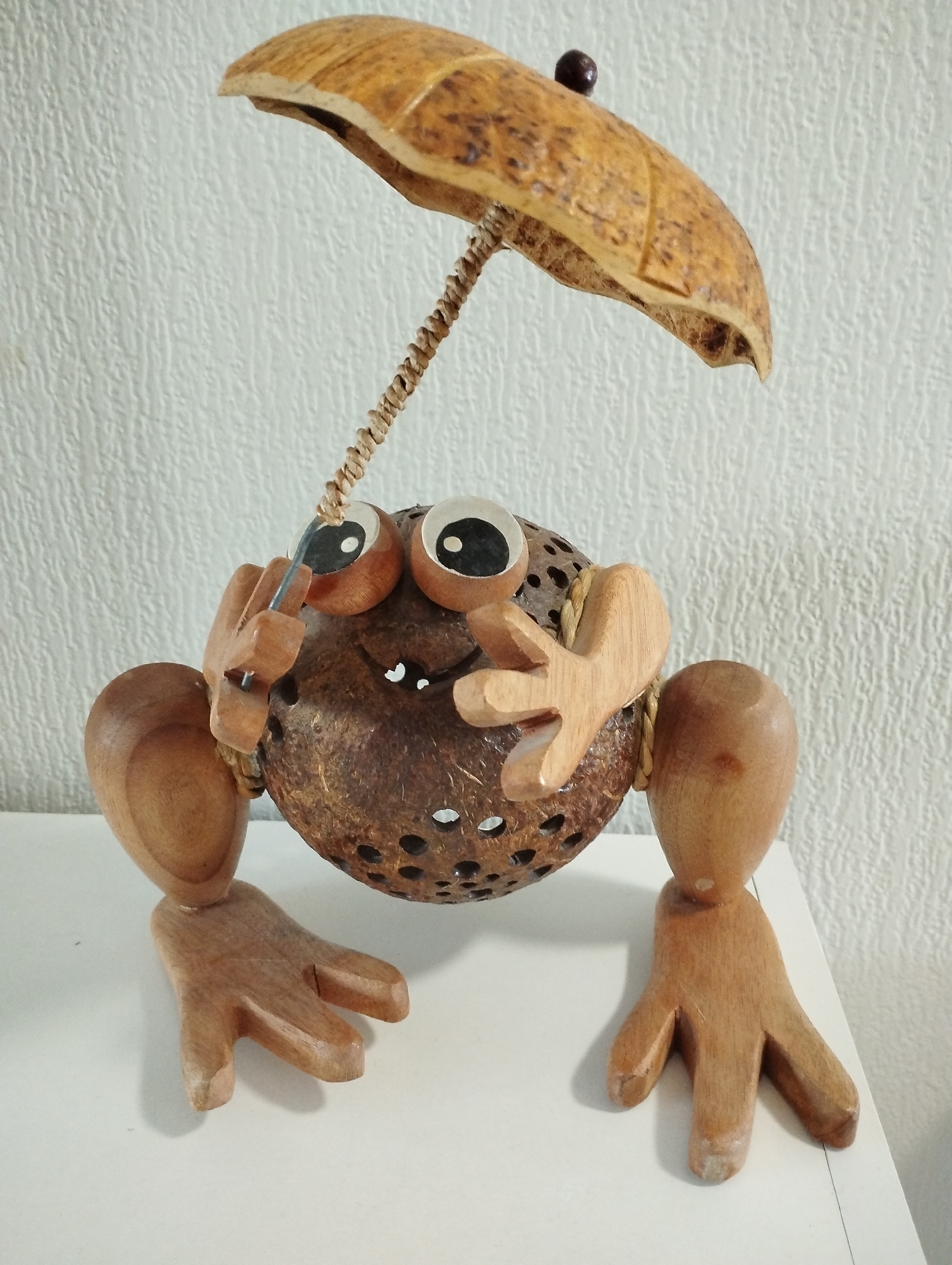 The image shows a handmade wooden frog figurine standing on a white surface against a textured, off-white wall. The frog's body is a coconut shell with small holes. It has large, googly eyes and wooden arms and legs connected to the body with rope. The frog holds a small, wooden umbrella made from a coconut shell above its head. The exact location in Sadovyy, Russia, cannot be determined from the image alone.