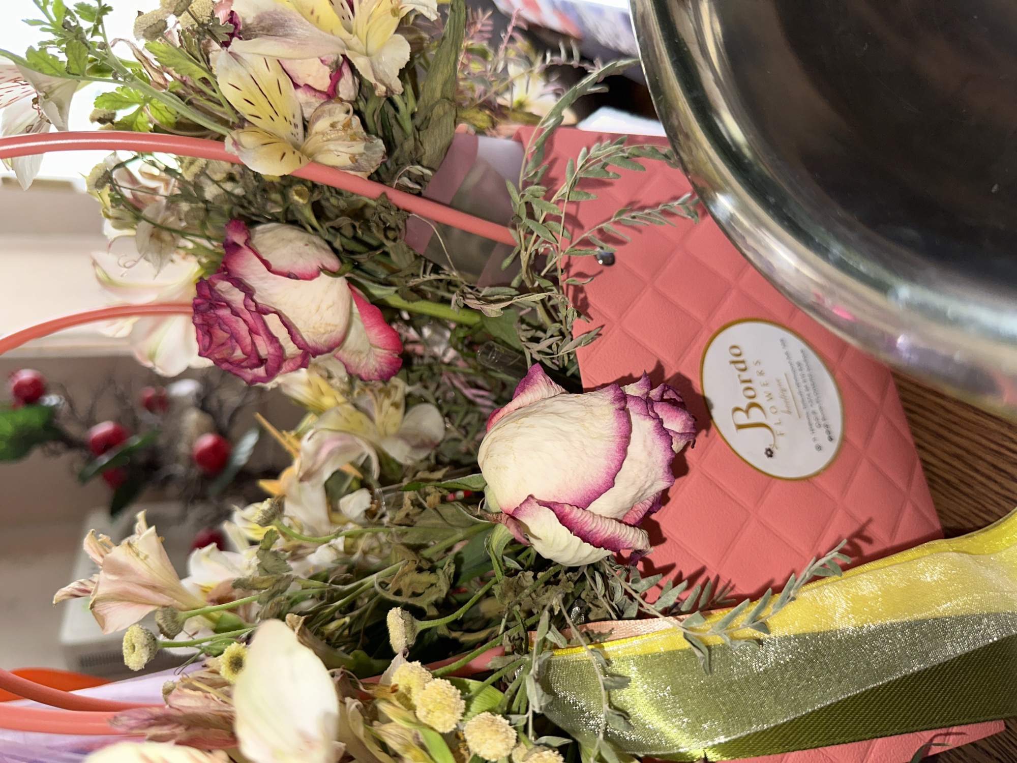 A bouquet of flowers is presented in a pink quilted basket. The basket has a textured surface and a quilted diamond pattern. The flowers appear to be a mix of roses and other blooms, some of which are wilting. There are also green ribbons tied around the handles of the basket. A label on the basket reads "Bordo Flowers boutique." The overall impression is that of a somewhat dried-out floral arrangement. The scene is set against a wooden surface, likely a table. In the background, there are hints of other objects and possibly a decorative branch with red elements. The lighting suggests an indoor setting.