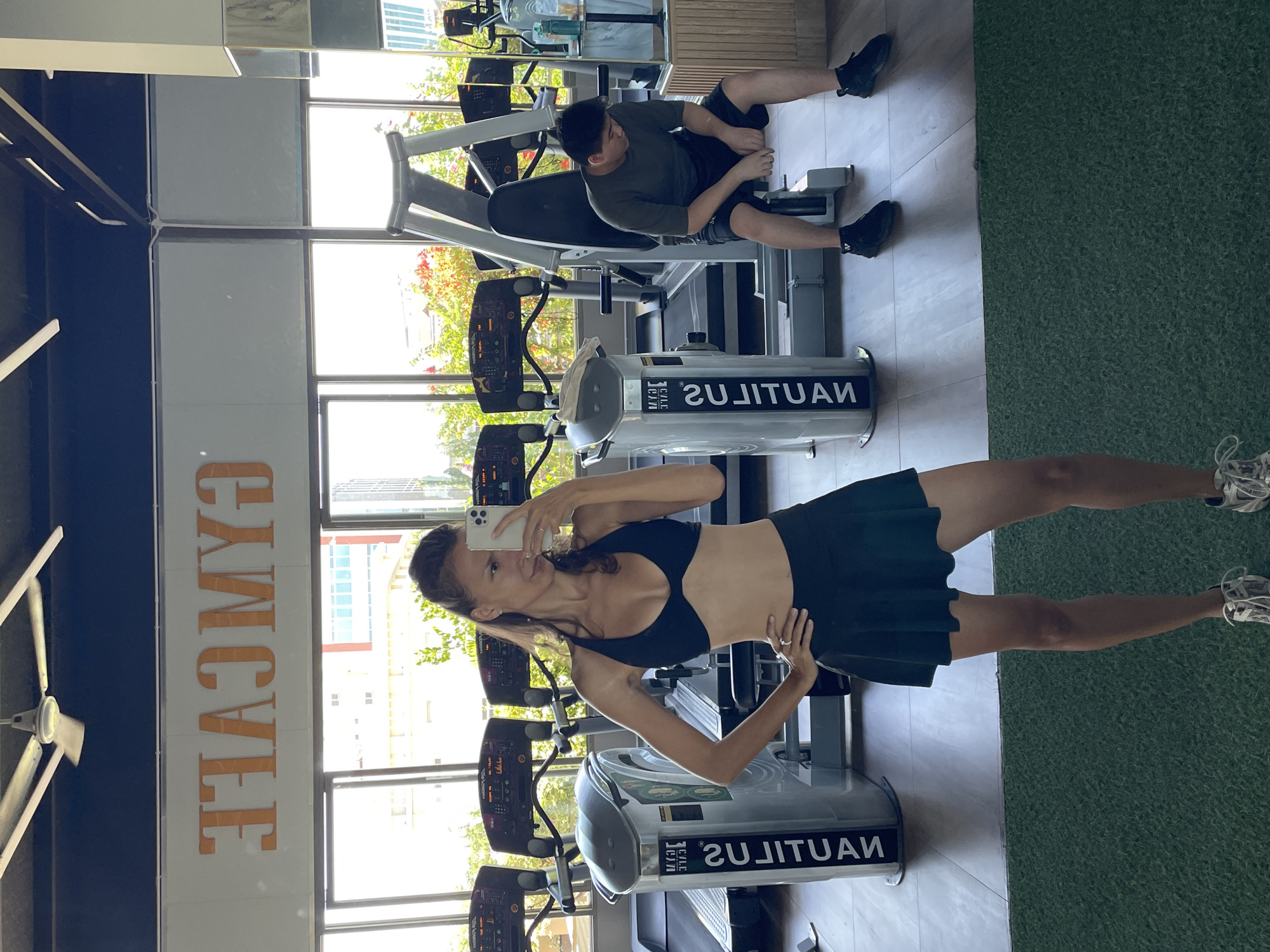 A woman is taking a mirror selfie in a gym. She is wearing a black sports bra and a black skirt. In the background, there are treadmills and a weight machine. A man is sitting on the weight machine, looking down. The gym appears to be located in Ham Thang Ward, Vietnam, as indicated by the "GYMCAFE" sign on the wall. The overall lighting suggests it is daytime.