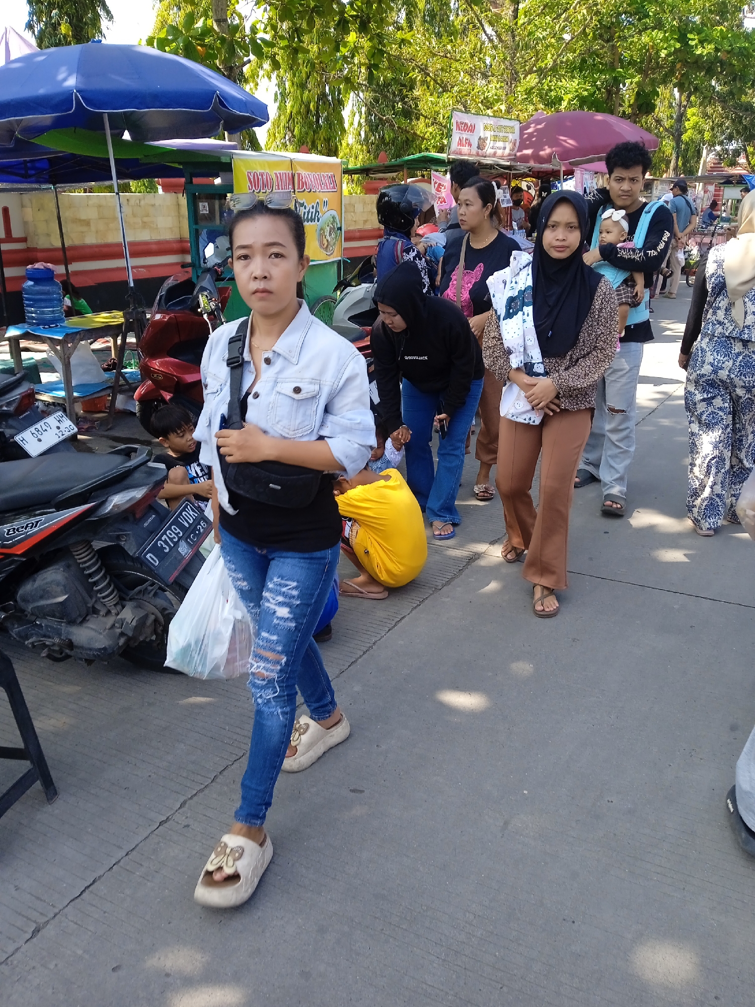The image captures a street scene in Kebondalem, Indonesia, on a bright, sunny day. A line of people is walking down the street, with a woman in a denim jacket, ripped jeans, and platform sandals in the foreground. She carries a black fanny pack across her chest and a white plastic bag in her hand. Beside her, partially obscured by a parked motorbike, a child is seen sitting behind the bike.
The line of people includes a woman wearing a hijab and holding a blanket, a man carrying a baby in a carrier, and other individuals walking in the same direction. Several food stalls are set up along the side of the road, identifiable by their colorful umbrellas and signage that says "SOTO AMIA BONOVLINE" and "SEKOND". The parked motorbikes indicate the area is accessible by vehicle. The environment appears to be a busy public space, likely a market or shopping area.