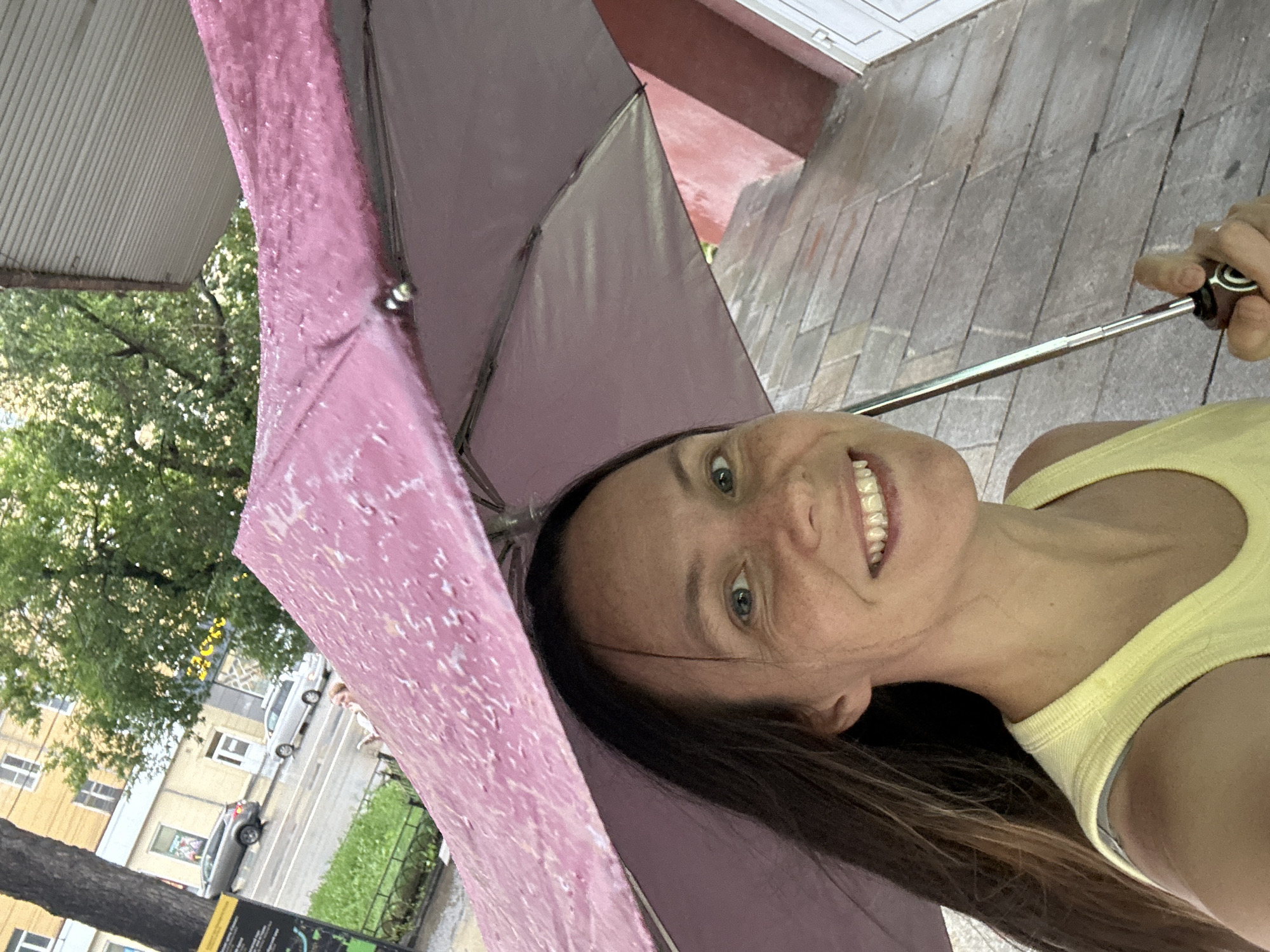 A woman is captured in a close-up, selfie-style shot, holding a pink umbrella over her head. She is looking at the camera with a wide smile, revealing her teeth. The umbrella, adorned with a speckled pattern, provides shelter from what appears to be rain, as evidenced by water droplets on its surface and the wet cobblestone pavement visible in the background. She is wearing a light yellow tank top. The background includes a building with a red facade, trees, and a street with cars, suggesting an urban setting. The overall scene conveys a cheerful mood despite the rainy weather.