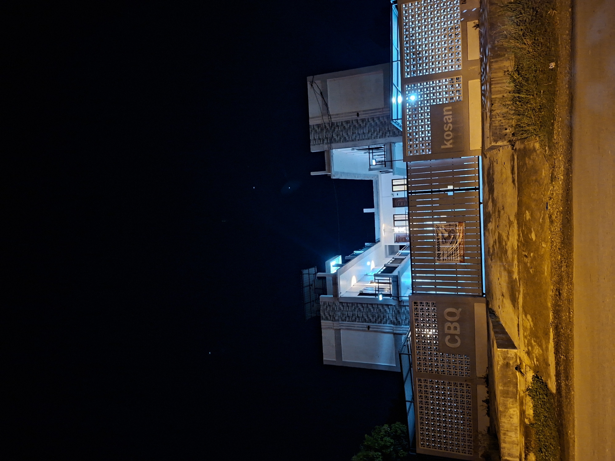 The image shows a building at night in Lhokseumawe, Indonesia. The building is two stories tall with modern architecture and blue lighting. The word "kosan" is visible on the right side, while "CBQ" is on the left. There is a gate in front of the building, along with a sidewalk and some vegetation. The sky is dark, with a few faint stars visible.