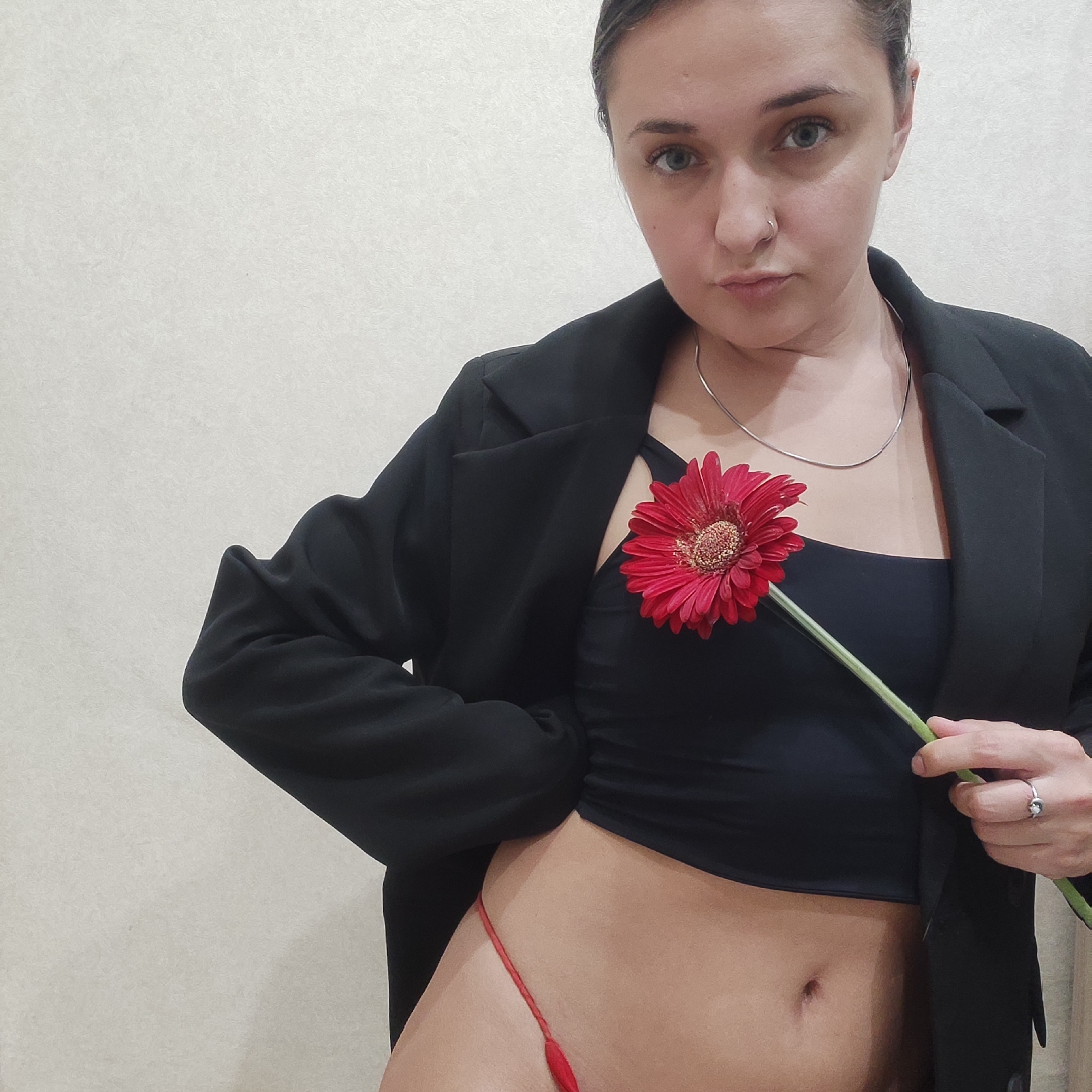 A woman is depicted in a medium shot against a plain off-white background. She is wearing a black blazer, open to reveal a black crop top underneath, and a red thong is visible. A red gerbera daisy is positioned against her chest, held by her right hand which also has a ring on the ring finger. She has a silver necklace and a nose ring. Her hair is pulled back. The location is unknown.