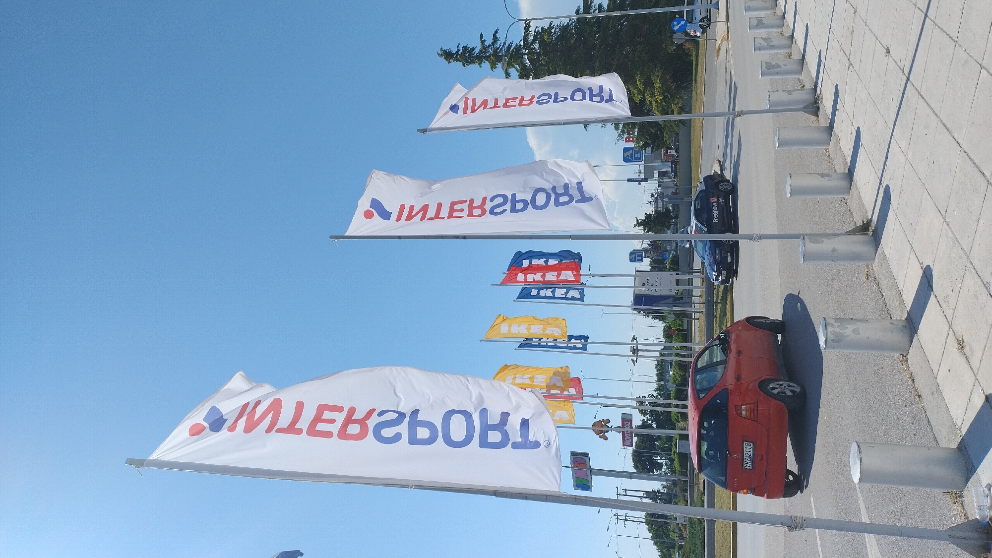 The image depicts a street scene in Pylaia Municipal Unit, Greece. The foreground features a sidewalk with several cylindrical metal bollards. A red Mercedes-Benz A-Class is parked on the left side of the street. In the background, multiple tall flags are displayed, with some featuring the Intersport logo and others displaying the IKEA logo in blue and red. A dark-colored Skoda with the "Freenow" logo is visible driving on the road. The sky is clear and blue, suggesting a sunny day. The environment appears to be an outdoor commercial area, likely near a shopping center or retail park.