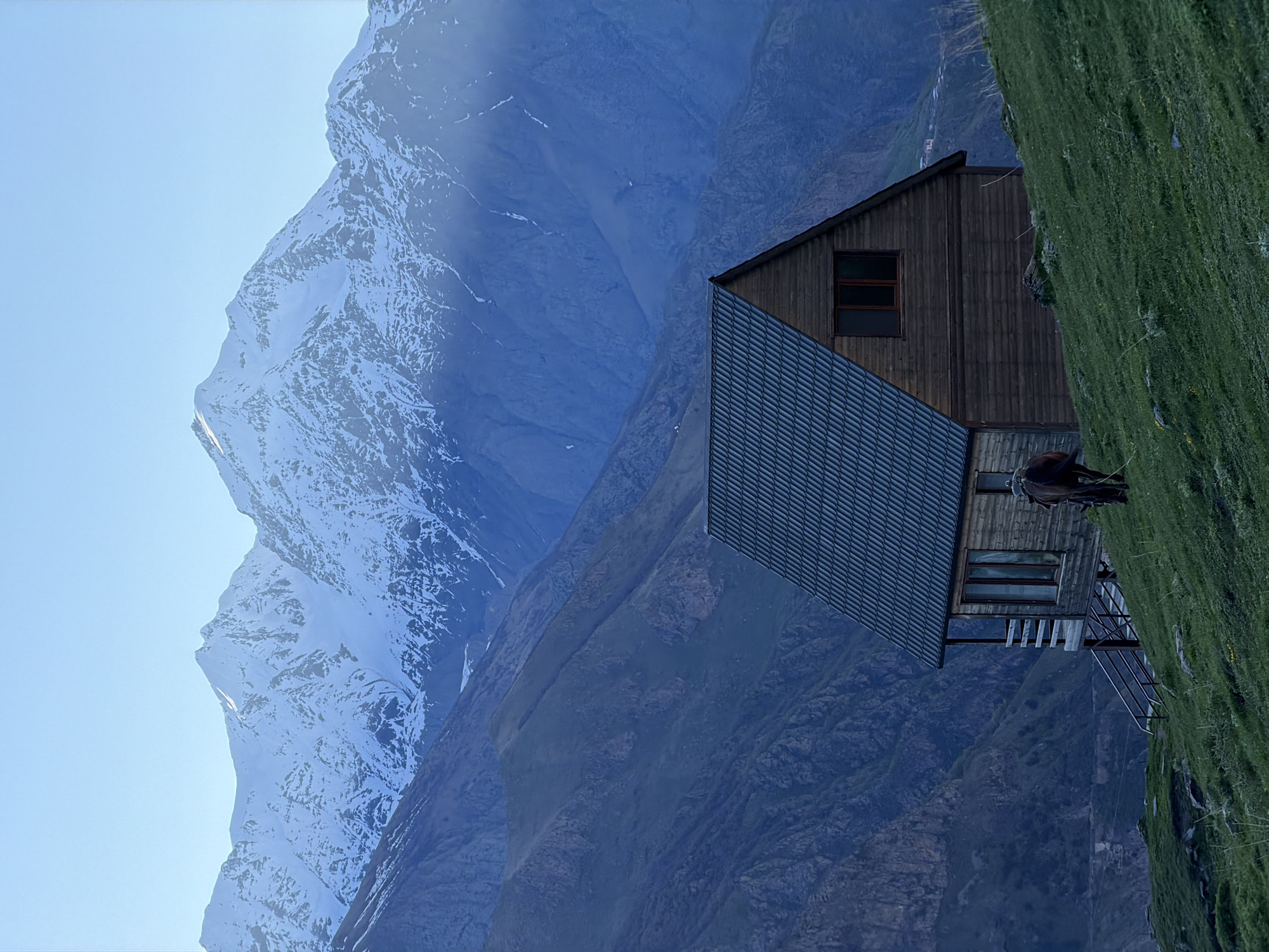 The image shows a building perched on a grassy slope in a mountainous environment. The building has a dark wood exterior, a corrugated metal roof, and several windows. A person and a horse are standing on the building's deck. In the background, a large mountain range is visible, with patches of snow on the peaks. The sky is clear and light blue. The location is Stepantsminda, Georgia.