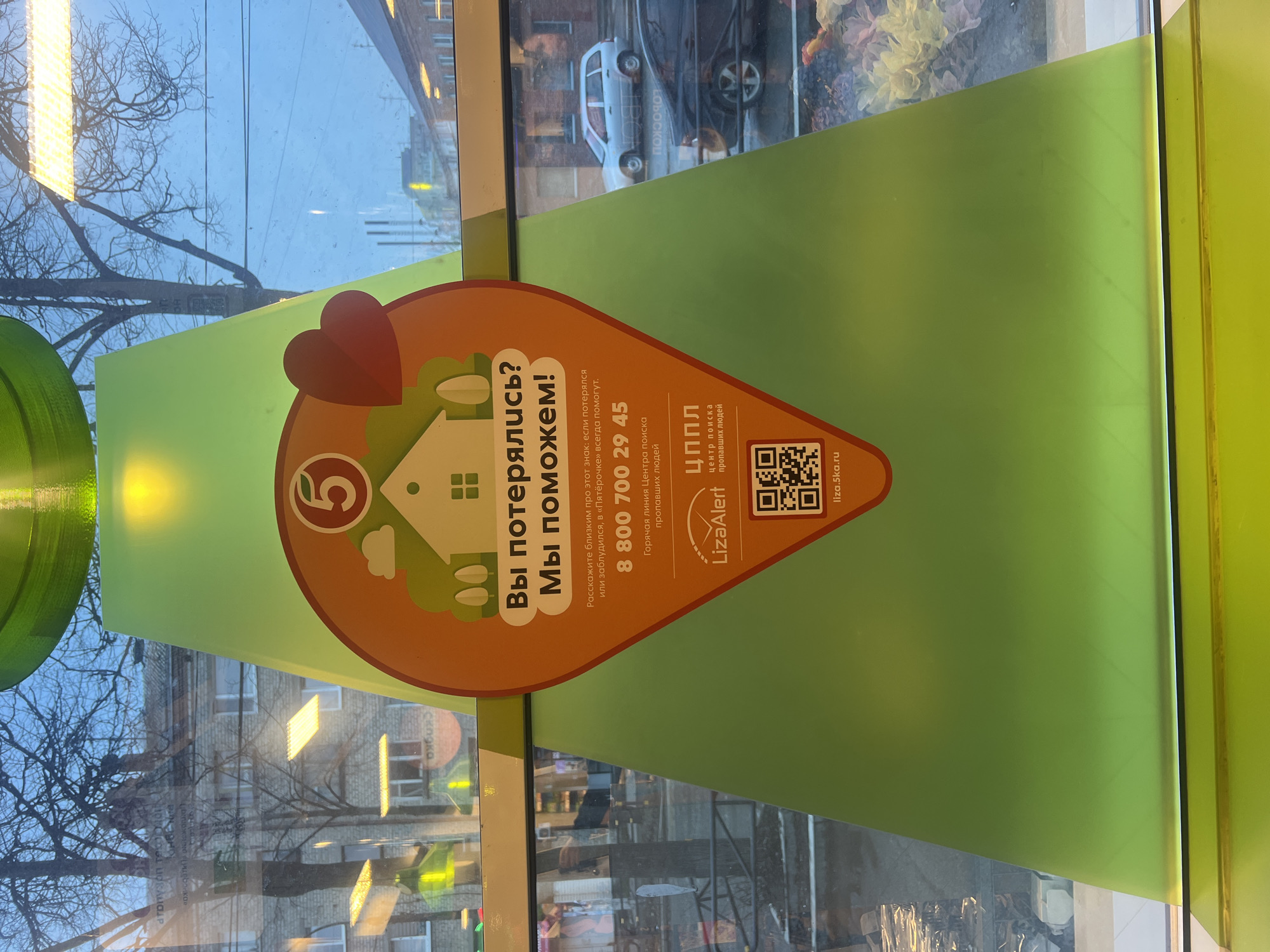 The image shows an orange and green sticker on a glass surface, possibly a window, with the words "Вы потерялись? Мы поможем!" ("Are you lost? We can help!") in Russian. The sticker also features a heart, a house, and the number 5 within a circle. Below this, it provides a phone number "8 800 700 29 45" and the website "liza.5ka.ru", suggesting it's a message from a search and rescue organization. The sticker mentions that in "Pyaterochka" stores, people can always help if someone gets lost or is disoriented. Outside the glass, a city scene is visible, including buildings, trees, and a glimpse of a car. The context suggests the location could be Chaikovsky, Russia, as the Cyrillic script indicates a Russian-speaking environment.