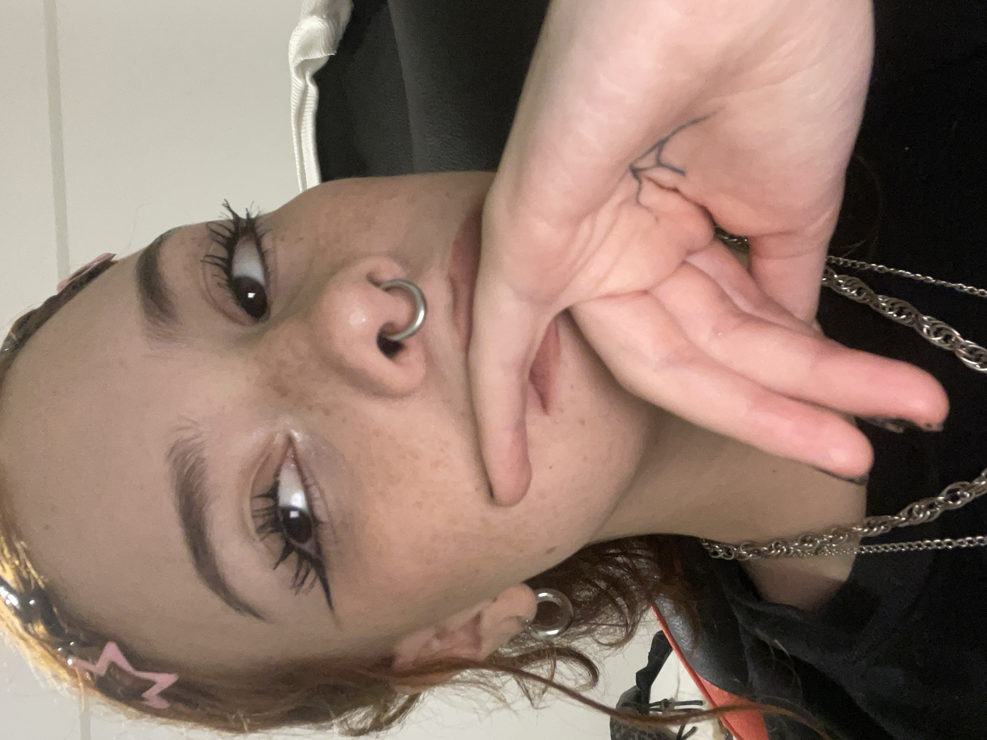 A close-up selfie of a fair-skinned person with freckles. The person has dark eyeliner, a nose ring, and what appears to be a hand tattoo. They are wearing multiple silver chains around their neck and have a hair accessory with pink stars. The background is out of focus, making it impossible to determine a location in Buenos Aires, Argentina.