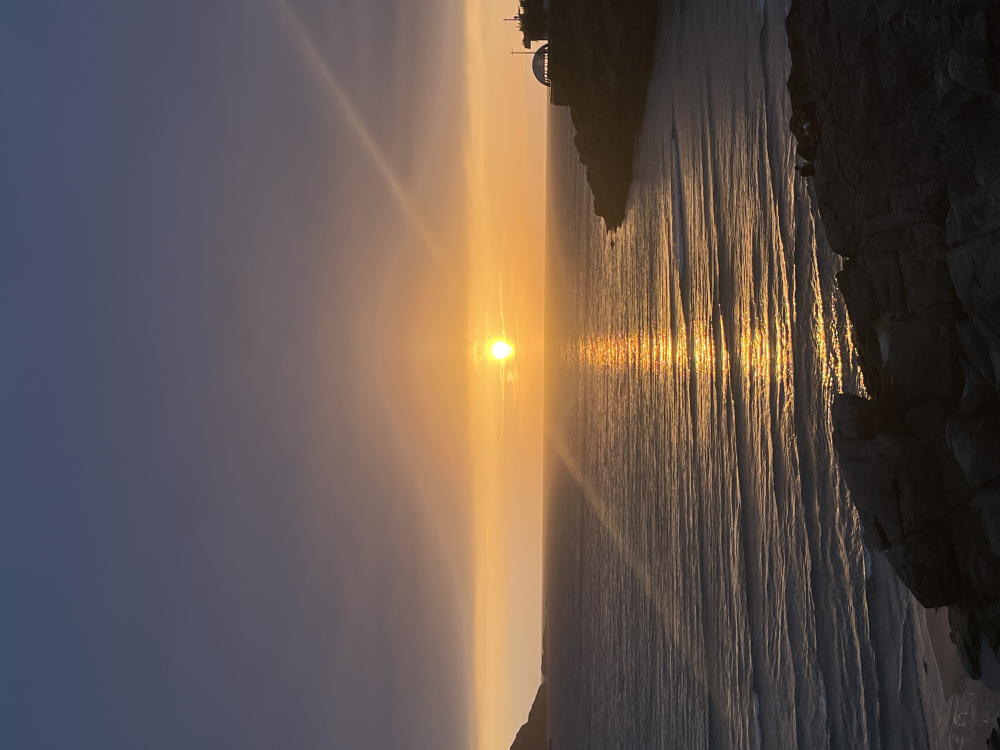The image shows a sunset over the Atlantic Ocean from A Coruña, Spain. The sun is a bright orb in the center of the frame, radiating light beams across the sky and reflecting on the water's surface as a vertical line of shimmering gold. The sky transitions from a pale blue on the left to a golden yellow towards the horizon. The coastline on the right is rocky and dark, with some structures visible on the top of a cliff. The water is rippled and reflects the golden light of the setting sun. There are no people visible.