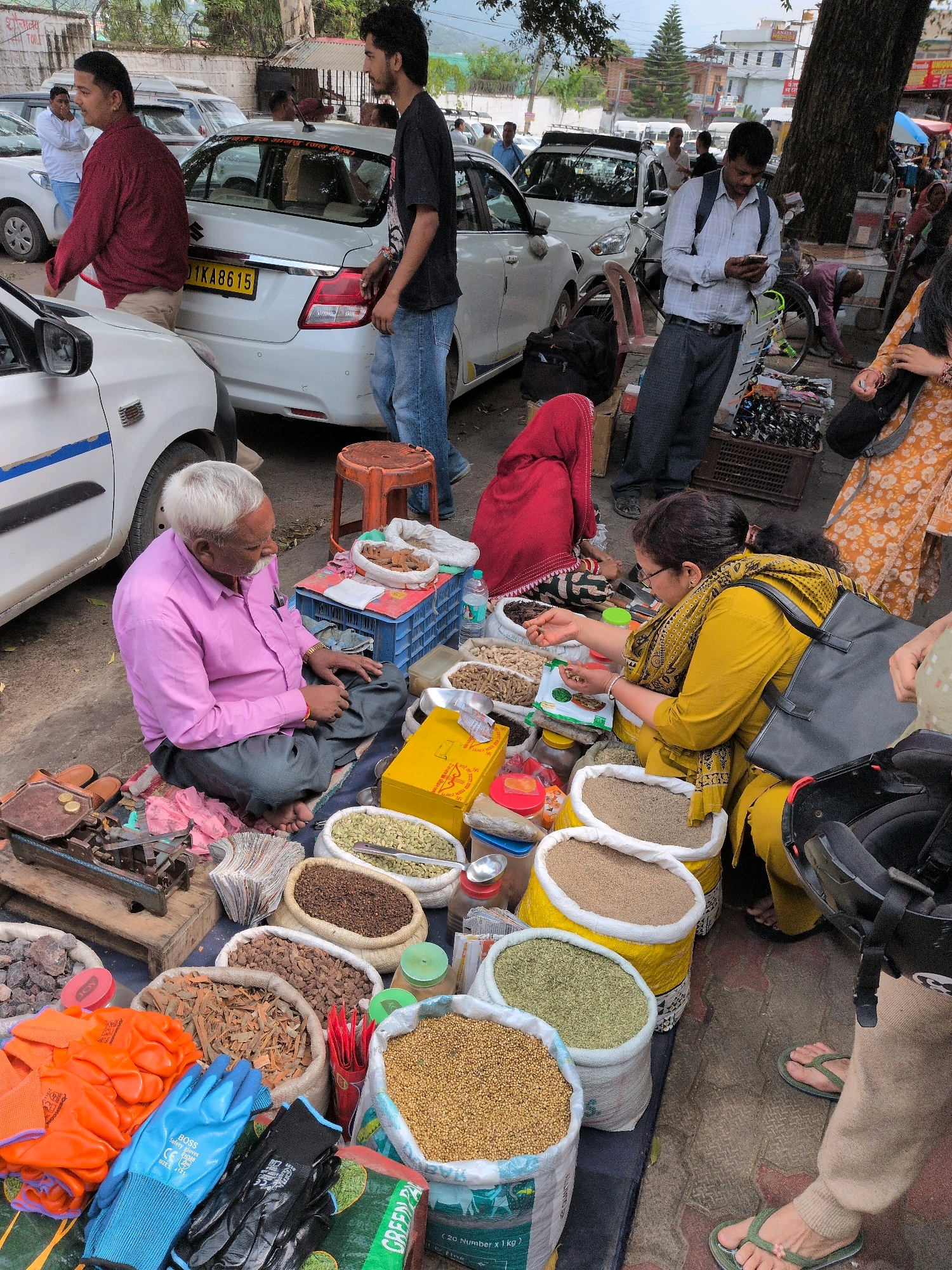 This image depicts a street scene in Bhuntar, India, likely a market area. The foreground is dominated by a vendor, an elderly man with white hair and a mustache, wearing a pink shirt and gray pants, seated cross-legged on the ground. He is surrounded by bags of spices and other goods for sale. To his right, another vendor, a woman wearing a yellow tunic and headscarf, is also seated and appears to be examining some items.
The merchandise includes various dried spices and grains in large sacks, such as what appears to be cardamom pods, coriander seeds, and other unidentifiable items. There are also smaller containers and bags of spices, a yellow box, and a set of orange and blue work gloves. A vintage weighing scale with coins on it sits on a wooden plank to the left of the seated elderly man.
In the background, several cars are parked along a street, including a white sedan with an Indian license plate. A few other individuals are visible, including men in casual attire and another person wearing a red head covering. The environment suggests a bustling roadside market, with buildings and trees visible further in the background. The overall impression is one of commerce and daily life in an Indian town.