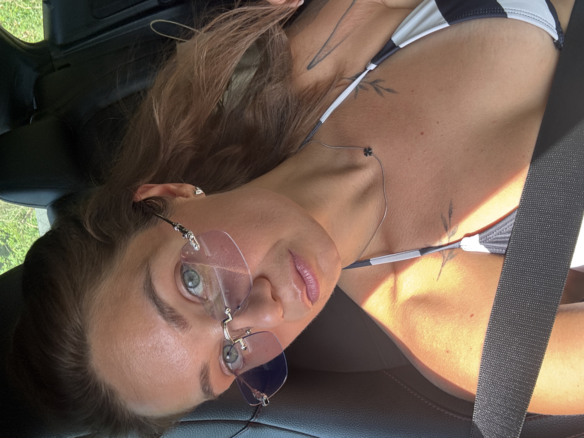 A female individual is depicted in a close-up selfie, seated in the front passenger seat of a vehicle in Sveti Stefan, Montenegro. She has medium-tan skin, light brown hair, and light blue-green eyes, and is looking directly at the camera. She wears rimless sunglasses with pink-tinted lenses, small heart-shaped stud earrings, and a thin silver necklace with a black four-leaf clover pendant. A black and white striped bikini top is visible, partially obscured by a dark seatbelt crossing her chest and left shoulder. Faint tattoos resembling leaf or branch motifs are present on her left clavicle/shoulder area. Her lips appear to have a glossy finish. Strong natural sunlight illuminates her right side, creating a diagonal shadow across her torso. The vehicle's interior features dark upholstery, and blurred green foliage is visible through the rear window.