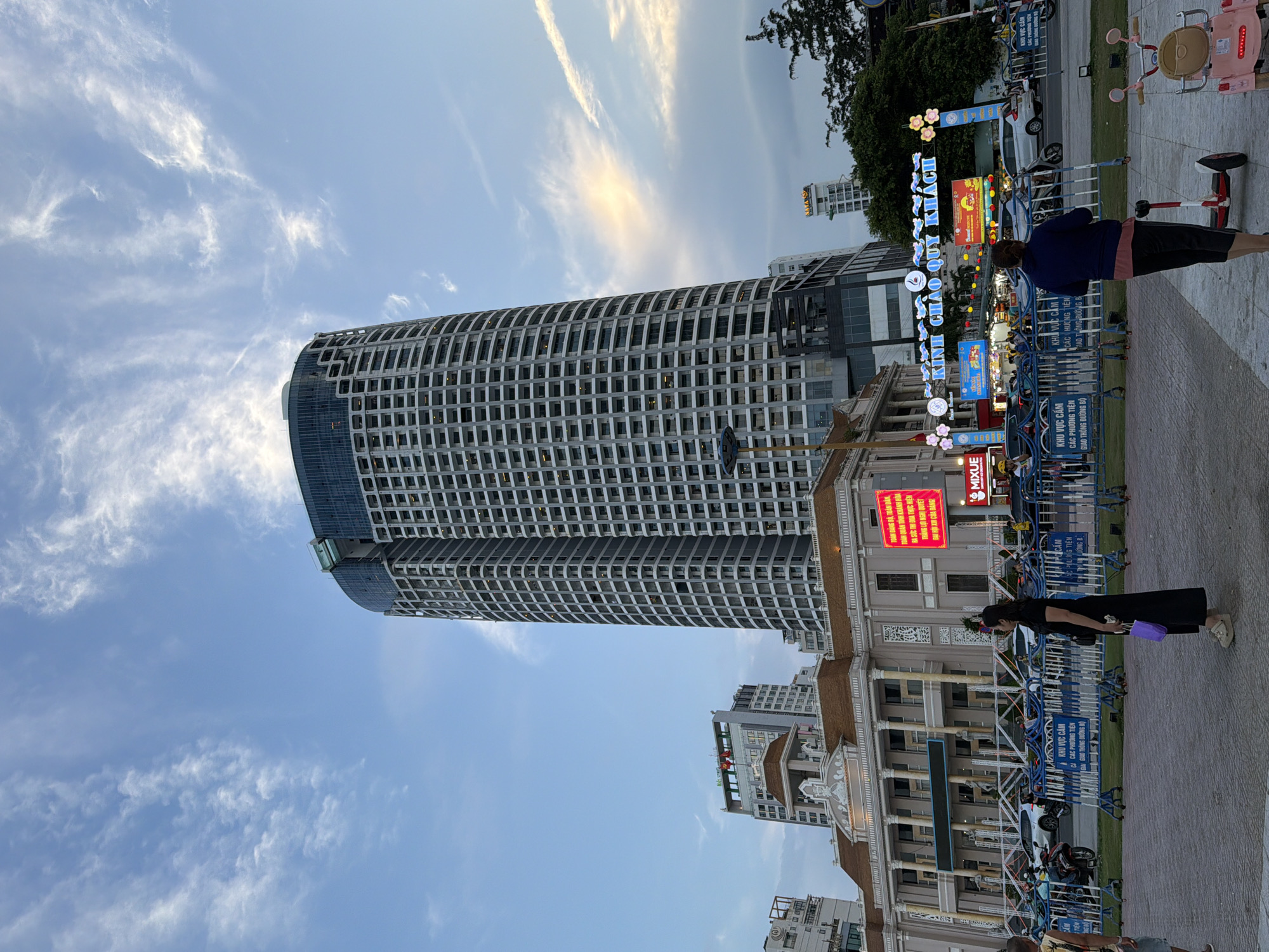The image depicts a street scene in Nha Trang Ward, Vietnam. In the background, a prominent, tall, curved skyscraper with a grid-like facade dominates the skyline, its upper section featuring darker glass. To the left of the skyscraper, a neoclassical-style building with a brown roof and white columns is visible. A large, illuminated LED screen displaying text in Vietnamese is mounted on this building's facade. In the mid-ground, an archway with blue lights and floral decorations spans across the street, bearing the text "KINH CHAO QUY KHACH". Below the archway, various signs and stalls are set up, with people and vehicles present. Blue barricades are positioned along the street, and cars and motorcycles are parked or moving. In the foreground, two individuals are standing on a paved area. One, on the left, is a person wearing a black dress and holding a purple bag. The other, on the right, is seen from behind, wearing a dark blue jacket and dark pants, with a small scooter-like device in front of them. A pink ride-on toy is also visible to the right. The sky above is a light blue with scattered, wispy clouds, and a hint of sunlight is breaking through the clouds on the right side. The scene appears to be set during the late afternoon or early evening.