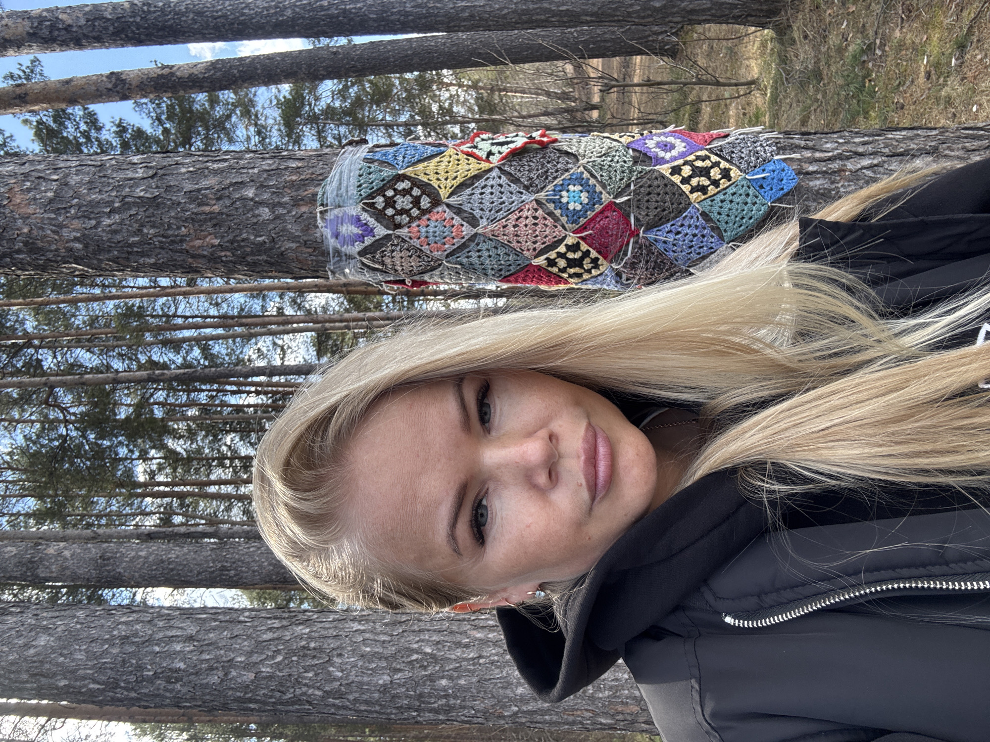 A portrait of a blonde woman with fair skin, wearing a black jacket, is framed against a backdrop of a pine forest. The woman's face is slightly tilted, and she is looking at the camera with a faint smile. Behind her, a felled tree trunk is visible, adorned with a crocheted quilt or blanket made of colorful granny squares. The forest consists of tall, slender pine trees with a clear blue sky visible through the branches. The location appears to be a natural environment, potentially a forest area within an urban setting. Unknown, Unknown.