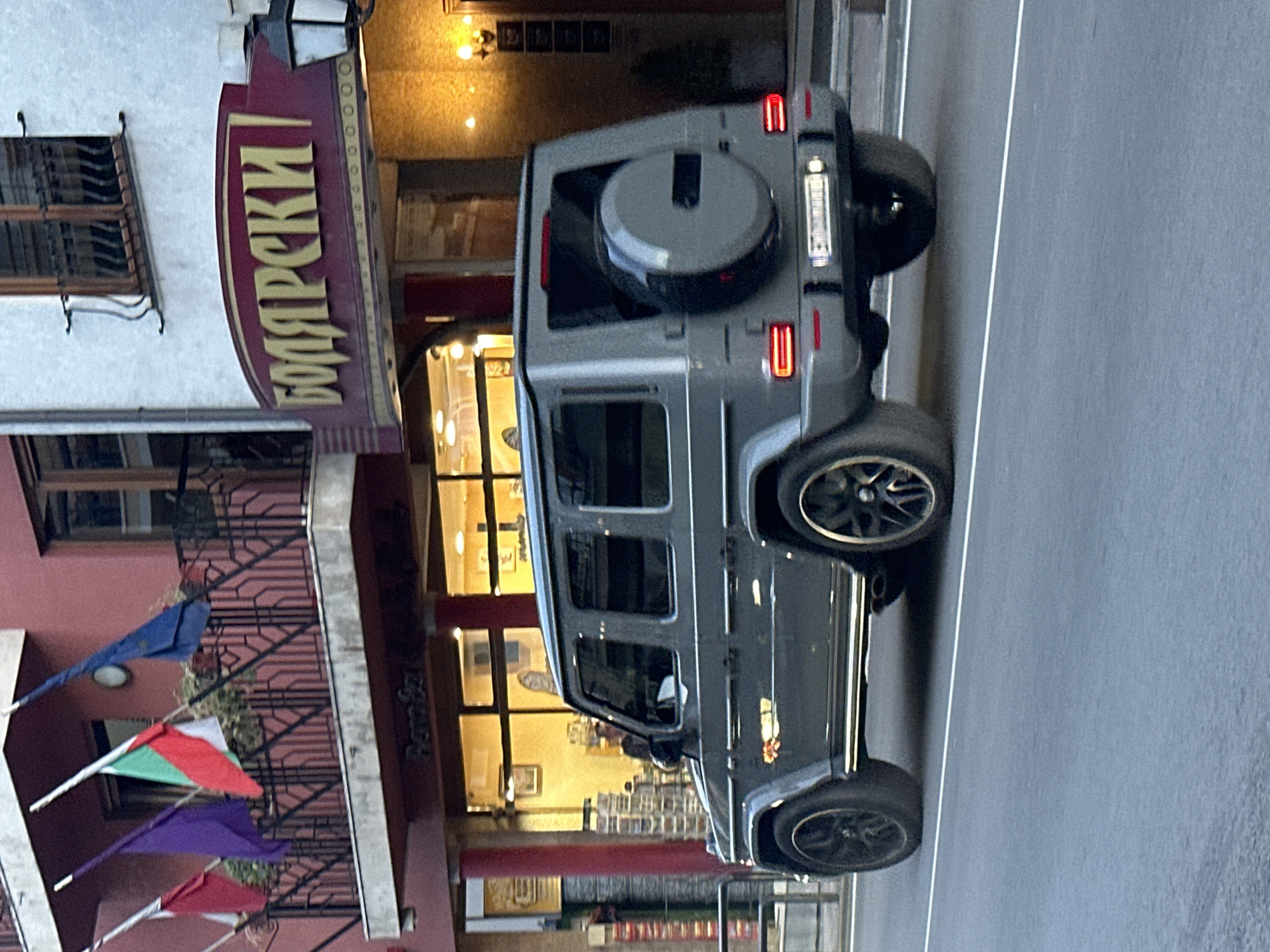 In Veliko Tarnovo, Bulgaria, a grey Mercedes-Benz G-Class SUV is observed driving on an asphalt road. The vehicle is facing away from the viewer, showcasing its rear and side profiles. The building behind the vehicle features a dark red facade with a white architectural element and a balcony adorned with flags, including the Bulgarian flag. Above a storefront, a large sign with Cyrillic lettering in gold appears to read "БОЛЯРСКИ". The storefront windows are illuminated with warm light, revealing glimpses of the interior. The sky is overcast, suggesting diffused lighting conditions.