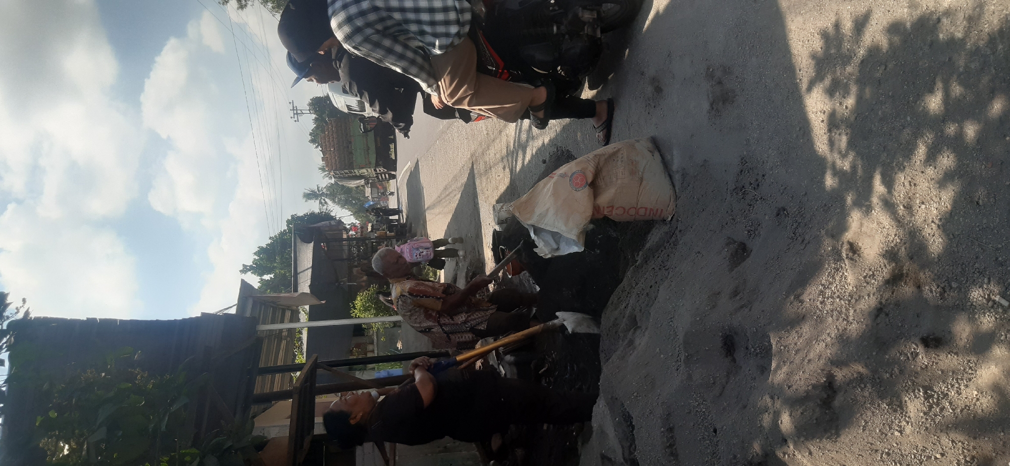 In Sepakek, Indonesia, a sunny day is depicted with a bright sky filled with scattered clouds. In the foreground, two individuals, an older woman in dark clothing and an older man in a patterned shirt, are actively engaged in mixing a dark substance with shovels on a dirt ground. Adjacent to them lies a large, light-colored bag with red lettering, likely containing construction materials, and a pile of sand. To the right, two people are seated on a motorcycle parked on the asphalt road. The road continues into the background, where a large truck carrying stacked materials is visible, along with other individuals walking. The scene appears to be in a semi-urban or rural area, with some structures and trees lining the roadside.