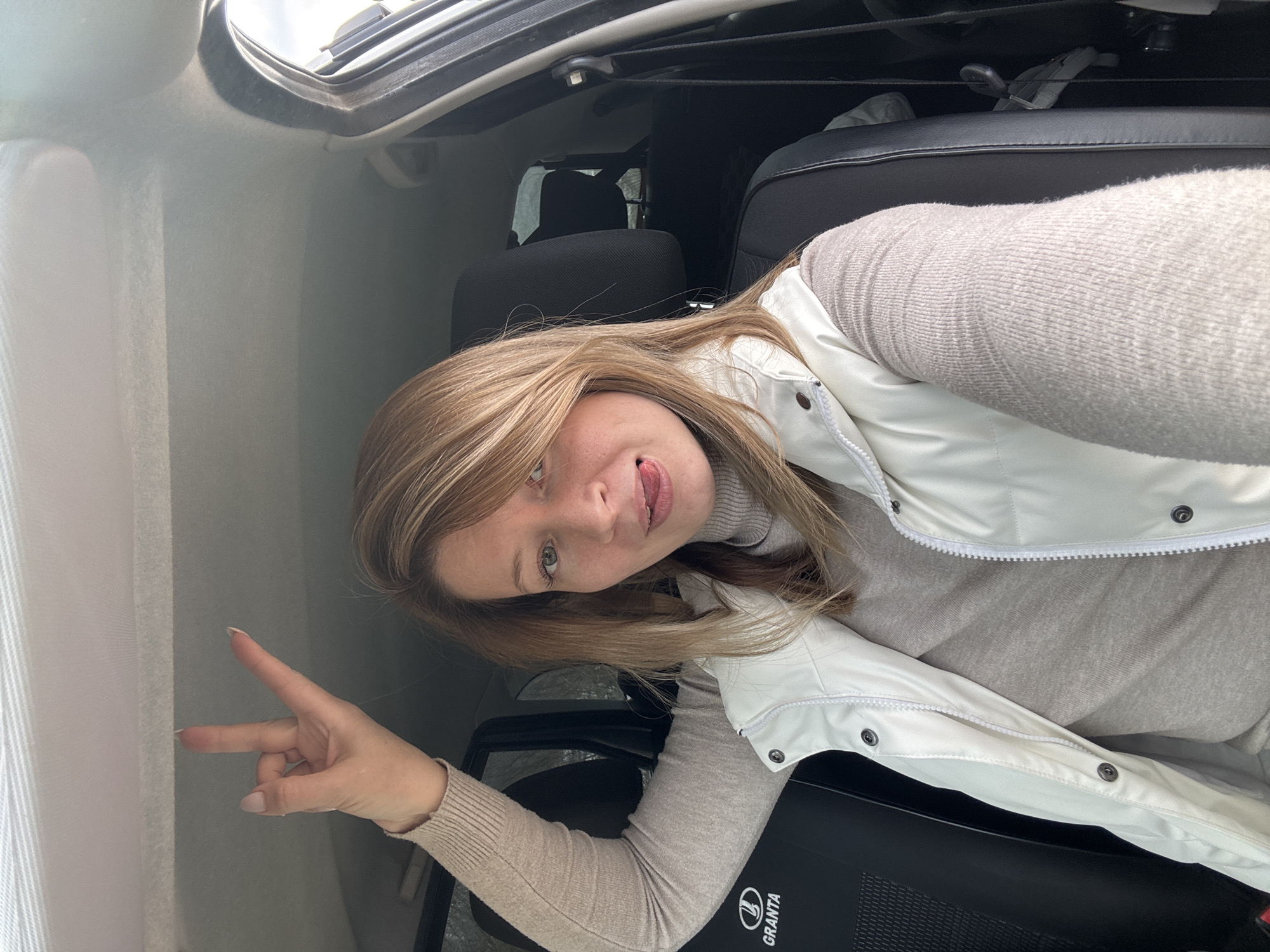 A woman is taking a selfie inside a car. She is blonde with light skin and is wearing a light beige turtleneck sweater and a white sleeveless jacket. She is sticking her tongue out and giving a peace sign with her left hand. Part of the car's interior is visible, including the roof, seats, and a "Lada Granta" logo on a black surface. The location is Вассята, Russia. The scene appears to be inside a parked vehicle.