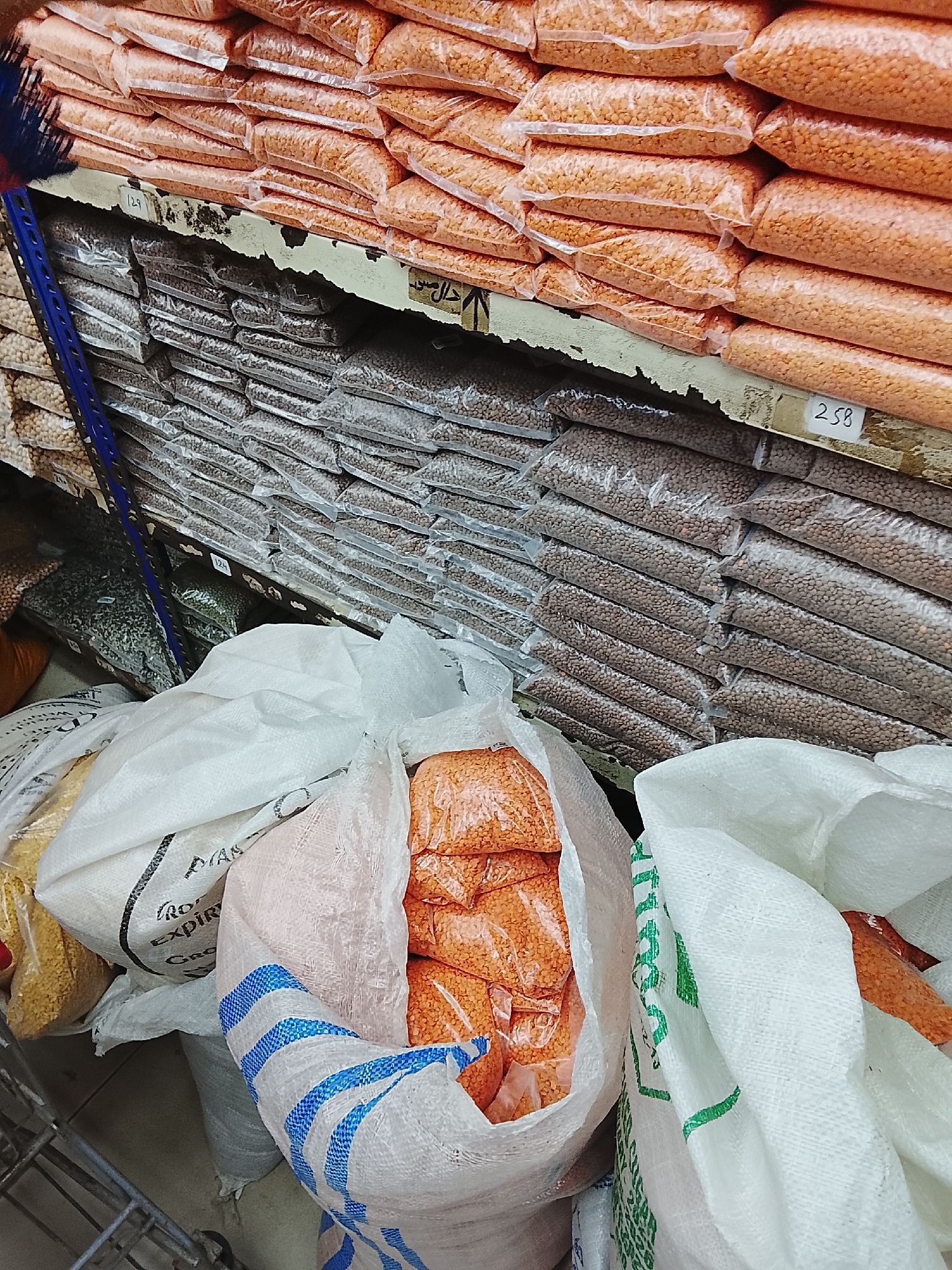 The image depicts a market scene in Gulgasht Colony, Pakistan, showcasing a variety of lentils packaged in both bulk sacks and smaller retail bags. The scene is densely packed, with shelves laden with numerous transparent bags of red and brown lentils. Below the shelves, large white sacks hold more lentils, including a bag partially open revealing smaller, sealed bags of red lentils inside. Additional bags of lentils in yellow and green are visible in the background. A blue and red broom leans against the shelving unit. A price tag labeled "258" is visible on the edge of the shelf.