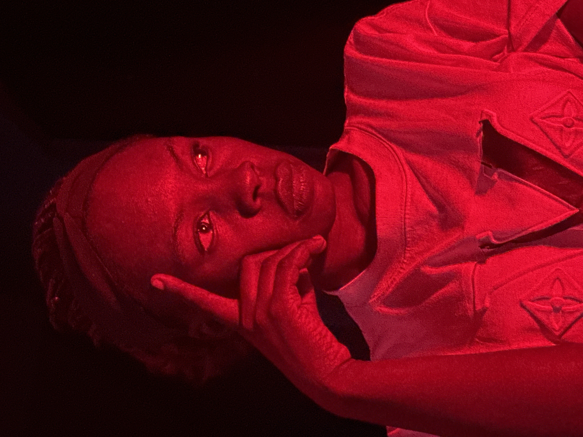 A medium shot captures a Black individual bathed in crimson light against a black background. The person, seen from the shoulders up, is looking to their right with their right index finger touching their temple. They are wearing a white t-shirt with a dark, embossed pattern on the chest, resembling a stylized 'V' within a floral motif. The lighting casts strong shadows, obscuring finer details of the environment. The scene's starkness and the color saturation suggest a controlled, possibly artistic, setting rather than an everyday outdoor or indoor location in Jos, Nigeria.