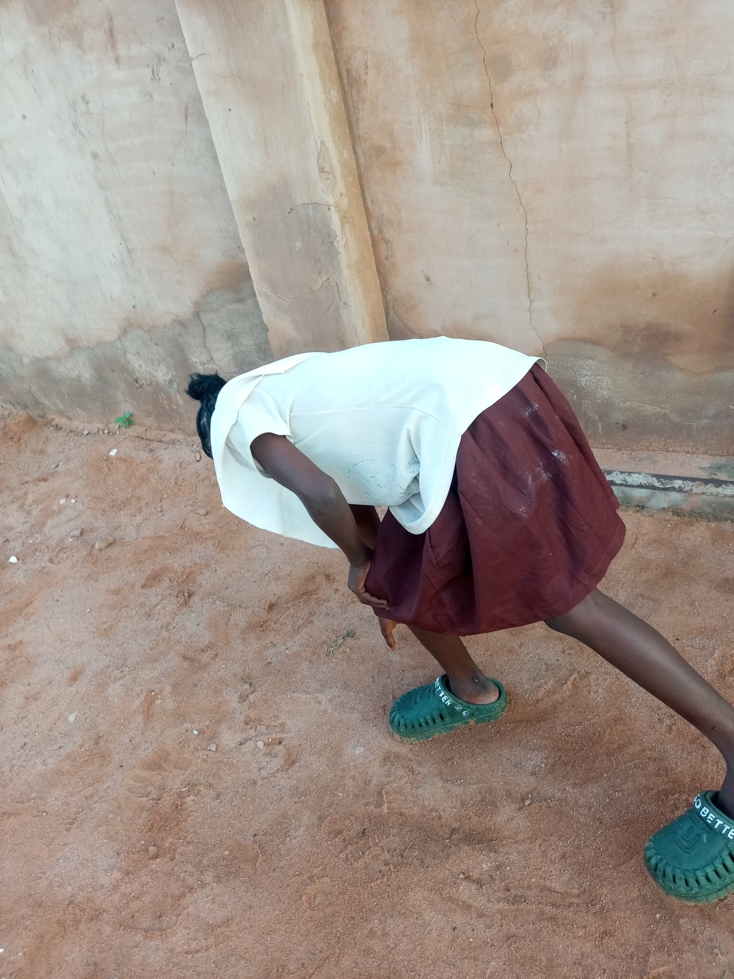 A young person, seen from a high angle and from behind, is bent over in a sandy area in front of a weathered, light-colored wall with visible cracks. The person wears a white short-sleeved shirt and a dark reddish-brown skirt. Their hair is pulled back into a bun. They are wearing green slip-on shoes, with the text "TO BETTER" visible on the side of the right shoe. The sandy ground has a few small pebbles and a small green sprout. The wall appears to be made of concrete or stucco and has various shades of beige and brown, with dark stains and rough textures. The scene is outdoors.