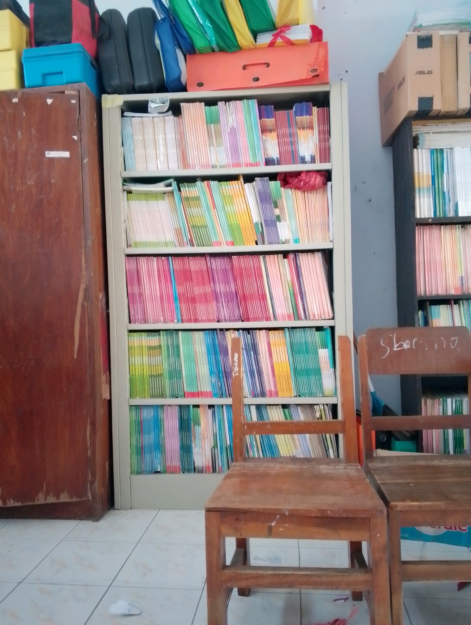 The image depicts a classroom or a storage area, likely in Papungan, Indonesia, characterized by an abundance of books and educational materials. In the foreground, two simple wooden chairs are positioned in front of a tall, light-colored metal bookshelf filled with rows of colorful books. To the left of the bookshelf is a tall, dark wooden cabinet with a single door. Behind the chairs and to the right, another darker bookshelf is visible, also packed with books. The walls appear to be painted a light blue-gray. On top of the cabinets and shelves, various items are stored, including what appear to be bags and boxes. The floor is tiled with a cream-colored grid pattern. A crumpled piece of paper is on the floor between the cabinet and the bookshelf. The lighting suggests an indoor environment, possibly during daytime.
