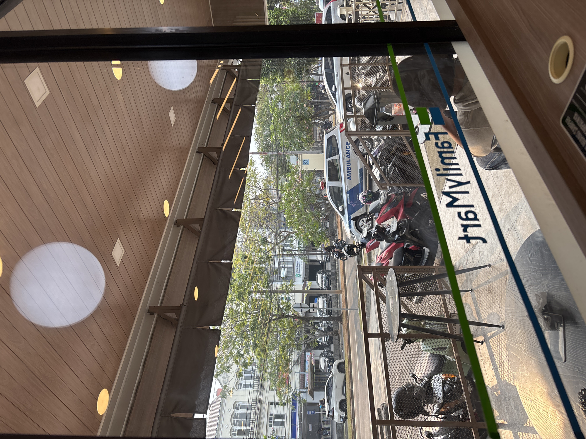 The image shows a street view in Binong, Indonesia, as seen through a window. In the foreground, the reflection of the interior of a room is visible, with a wooden ceiling and circular light fixtures. A person with dark hair is seated and looking away from the camera. In the middle ground, parked motorcycles are visible behind a fence. A round table with three legs is also present. Beyond the fence, a street runs horizontally across the image. Several cars and an ambulance are parked along the street. Buildings line the opposite side of the street, with trees and foliage in between. The sky is bright and appears to be daytime.