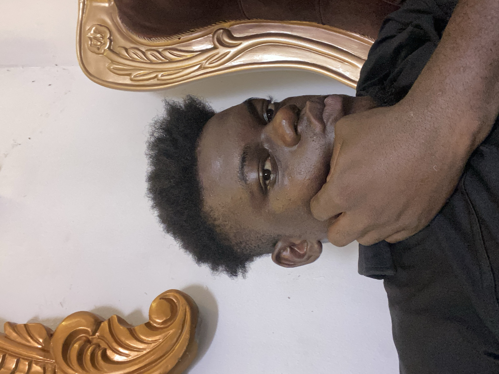 An image, rotated 90 degrees clockwise, features a young person with dark skin and short, curly dark hair. The person is looking directly at the viewer with an open-eyed gaze. Their right hand is positioned over their chin and mouth area, partially obscuring the lower face, which could suggest a contemplative or pensive mood. They are wearing a dark-colored, likely black, garment, possibly a t-shirt or shirt.
The setting appears to be indoors, against a plain white wall. Flanking the person's head and body, both above and below in the current orientation, are sections of ornate, golden-colored furniture. These pieces have intricate carvings and a glossy finish, suggesting decorative elements of a chair, sofa, or headboard. The overall lighting is consistent with an indoor environment. There is no visible text in the image.
