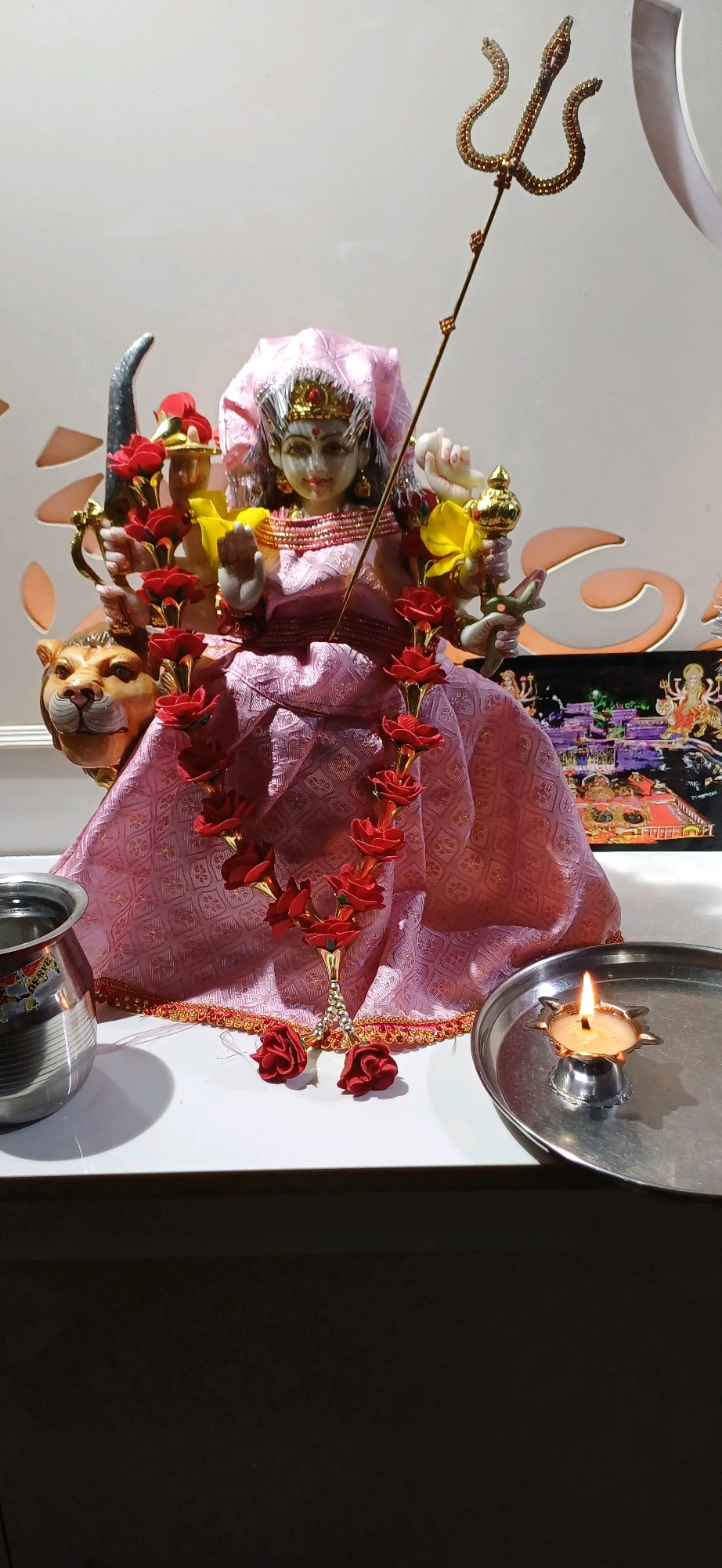 This image depicts a religious altar, likely in Ajmer, India, dedicated to the Hindu goddess Durga. The central subject is a statue of Durga, adorned in a pink and gold-patterned sari and a pink veil. She is surrounded by decorative red rose garlands and holds a golden trident, a symbol of her divine power. To her left, a statue of a lion, her vahana (mount), is visible. In the foreground, a small lit diya (oil lamp) rests on a silver plate, signifying worship and spiritual offering. A stainless steel pot, possibly containing water for rituals, is placed to the left of the altar. The background features a patterned wall and a framed image depicting other Hindu deities, suggesting a devotional space within a home or a temple. The lighting is warm and inviting, highlighting the sacred objects and creating an atmosphere of reverence.