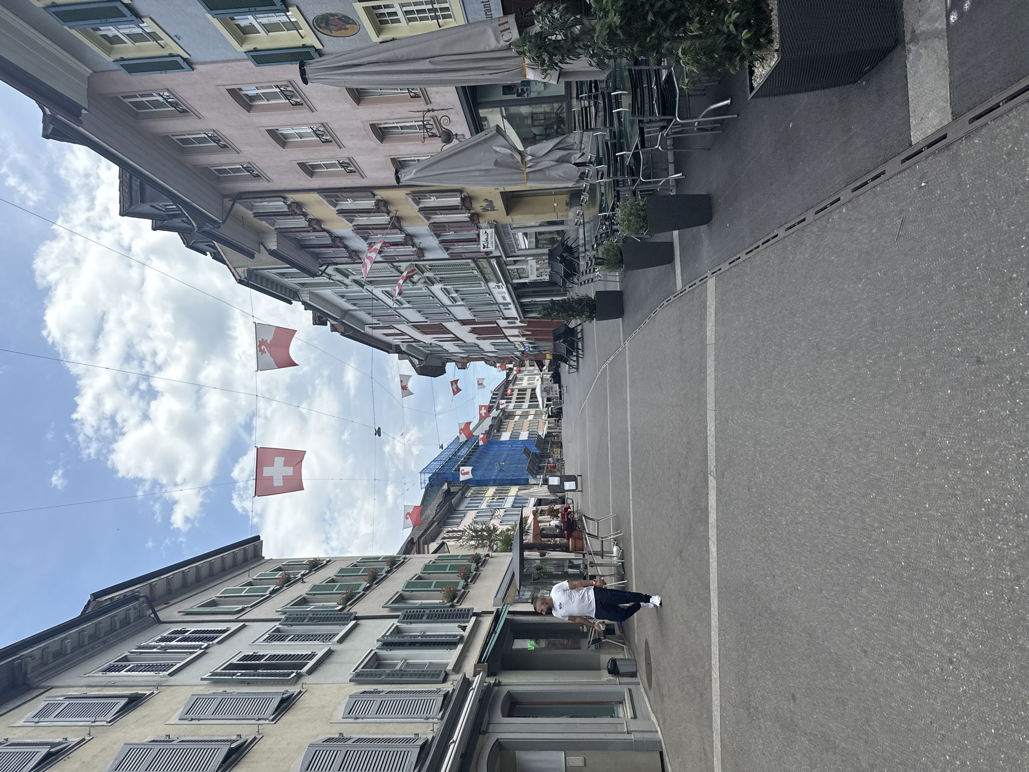The image shows a street scene in Liestal, Switzerland. The view is looking down a narrow street flanked by buildings on both sides. Buildings have multiple stories and diverse architectural styles, some with window shutters and flower boxes. Swiss flags are strung across the street. A man wearing a white shirt and dark pants is walking along the right side of the street. Tables and chairs are set out along the building fronts, suggesting outdoor dining areas. The sky is visible between the buildings, displaying white clouds against a blue sky. A building is under construction, being wrapped in blue material.