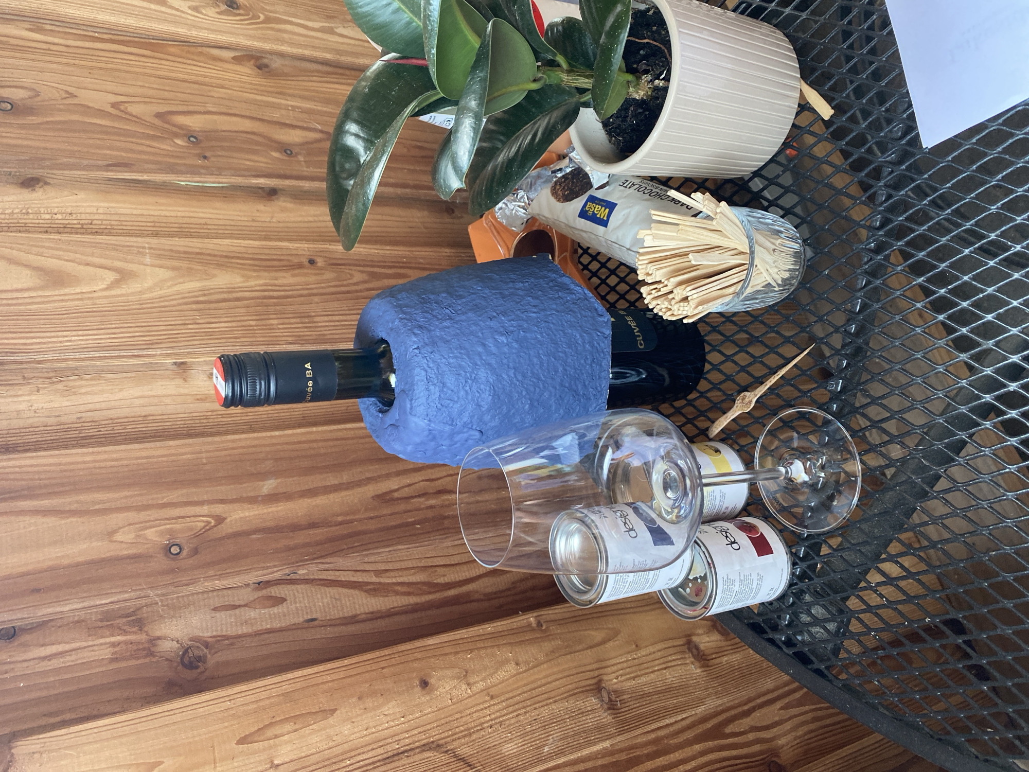 An outdoor table with a metal mesh surface displays various items against a backdrop of wooden planks. On the table, a bottle of wine is partially covered by a blue, textured material. To the left of the wine bottle, a wine glass stands over two small cans, one labeled "design". A small potted plant with large green leaves is positioned to the right of the wine bottle. A small glass container filled with wooden stir sticks is in front of the plant. A few cookies in a foil wrapper are visible behind the stir sticks. A single piece of white paper rests on the table in the foreground. The scene appears to be set up for a casual gathering or activity. The image does not provide any information that would allow for its location to be determined as Lütjenwestedt, Germany.
