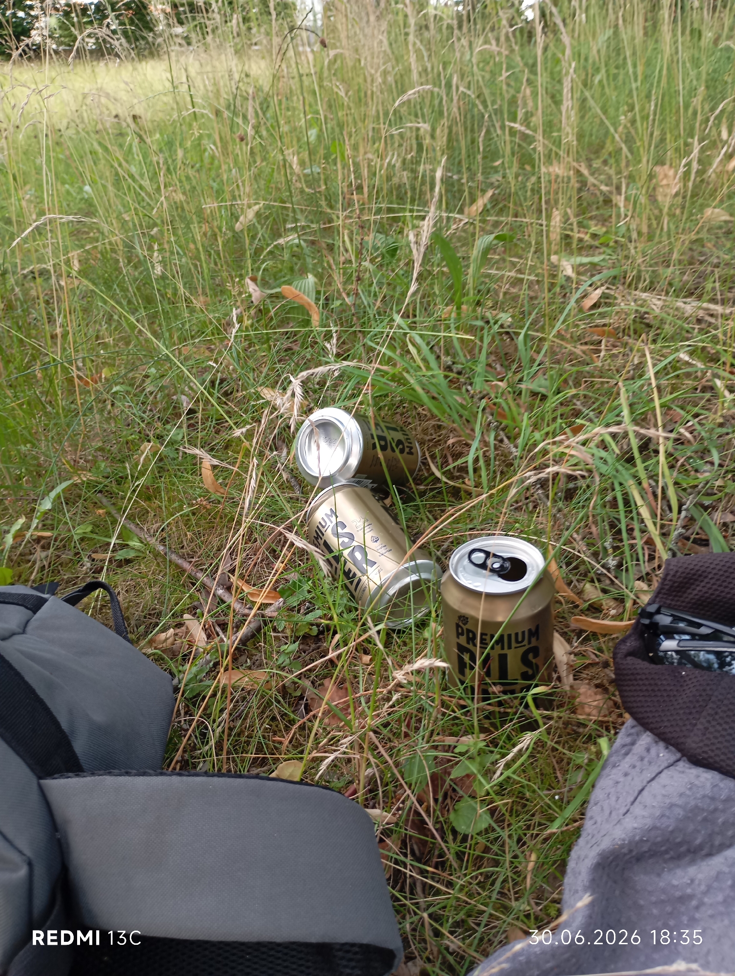 The image shows a ground-level view of a grassy area with several empty beer cans scattered among the tall grass and dry leaves. A gray backpack is visible in the lower left corner, partially obscuring some of the cans. To the right, a dark cloth, possibly a jacket or blanket, is laid out, with a pair of sunglasses resting on it. The scene suggests a casual outdoor gathering or a break taken during an outing. The lighting indicates it is daytime, likely late afternoon given the timestamp. The timestamp "30.06.2026 18:35" and the text "REDMI 13C" are visible at the bottom of the image. The overall impression is one of relaxation and perhaps a bit of littering, as the discarded cans are not in a trash receptacle. The setting appears to be a natural, undeveloped area, with tall grasses and some sparse trees in the blurred background. There are no people visible in the frame.
