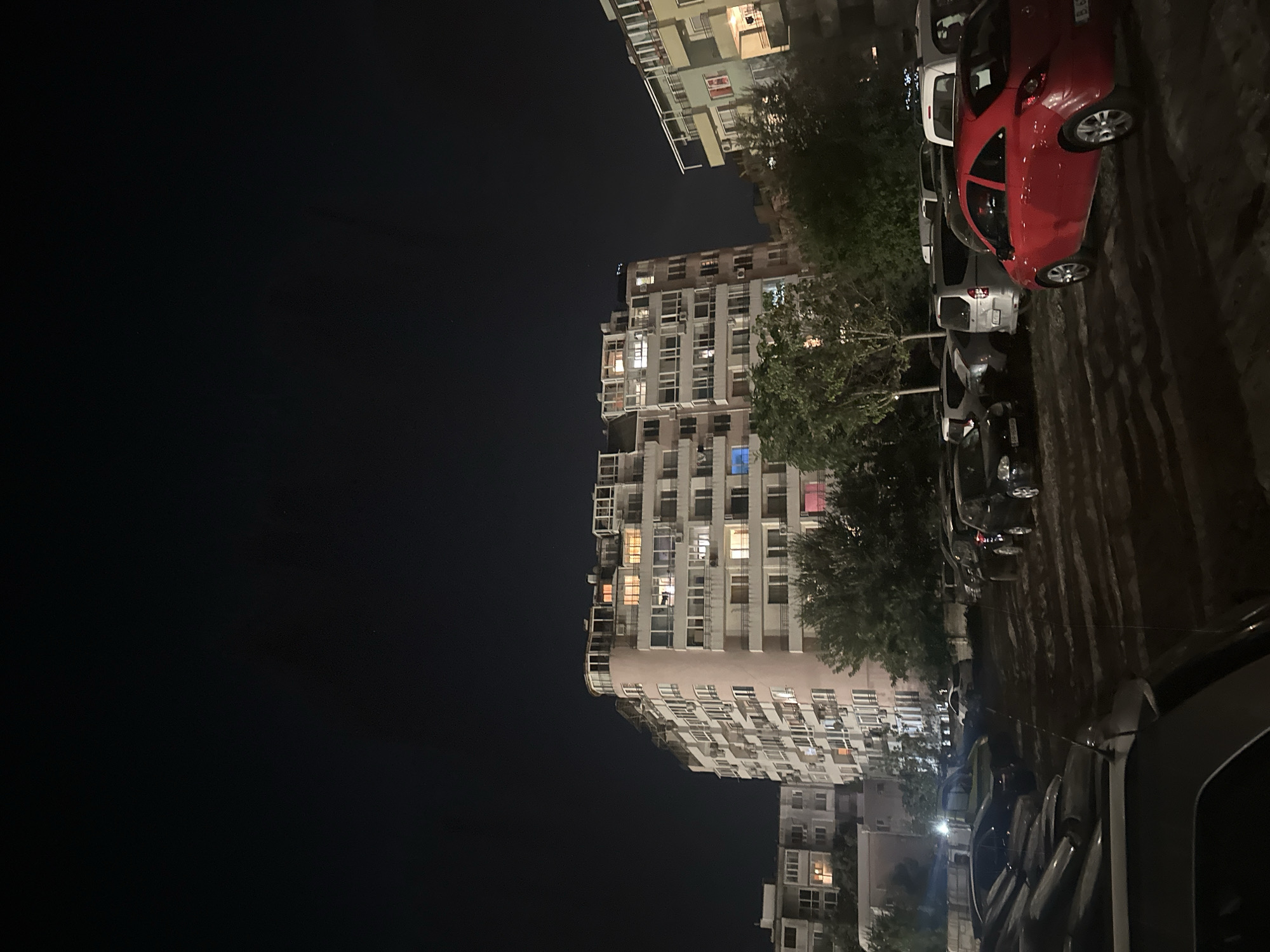 The image shows a multi-story apartment building at night, with lights on in many of the windows. In front of the building is a parking lot filled with cars. A red car is prominently parked in the foreground. Trees and other buildings are also visible in the background. The overall scene suggests a residential area at night.