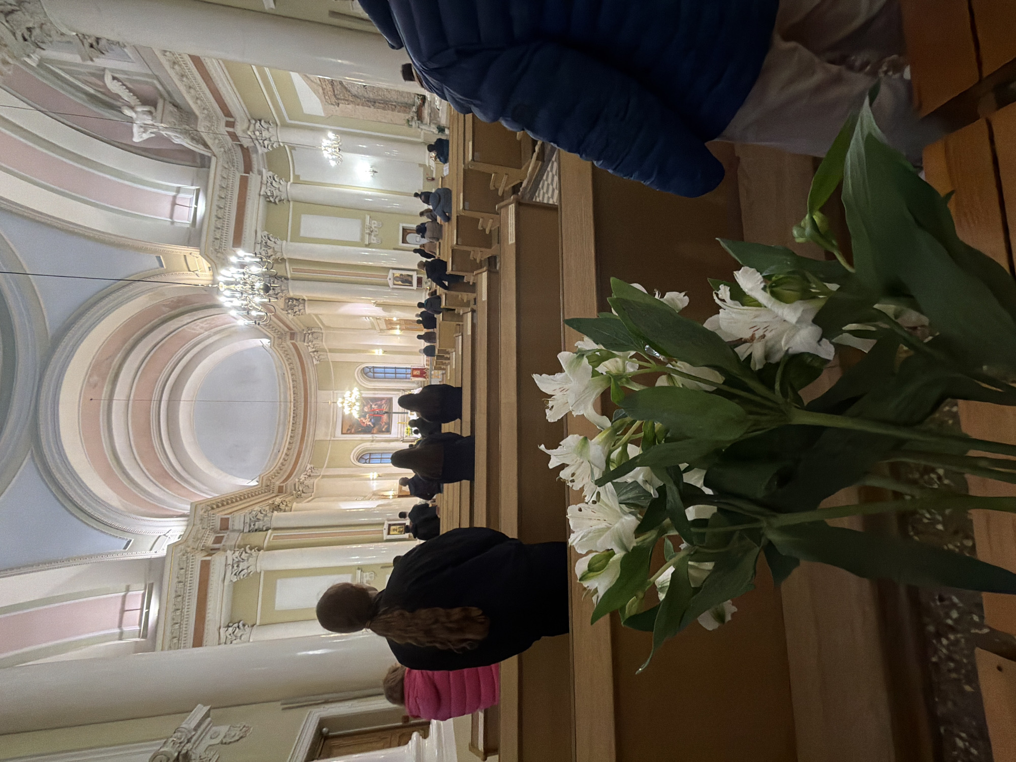 The image depicts the interior of a grand church or cathedral, viewed from a pew. The perspective is looking forward towards the altar area, with a significant portion of the high, ornate ceiling and upper architecture visible.
In the immediate foreground, to the right, is a prominent bouquet of white flowers with green leaves, possibly lilies or alstroemerias. To the left of the flowers and extending into the mid-ground, rows of wooden pews are visible, occupied by several individuals. Most people are seen from behind, seated or kneeling reverently, facing the front of the church. Their attire is mostly dark, with a person in a blue puffer jacket visible in the foreground right, and another in a pink top further back.
The church interior is characterized by classical or Baroque architectural elements, featuring tall columns, intricately decorated cornices, arched ceilings, and decorative moldings in shades of cream, pastel pink, and light blue. Multiple ornate chandeliers hang from the ceiling, illuminating the space with a warm glow. In the distance, towards the front of the church, an altar area is visible, adorned with paintings or icons. Another large painting can be seen on the upper wall to the left.
The overall atmosphere suggests a religious service or a moment of quiet prayer. No visible text is legible in the image due to distance and resolution. The lighting appears artificial, suggesting it might be evening or a dimly lit day indoors.