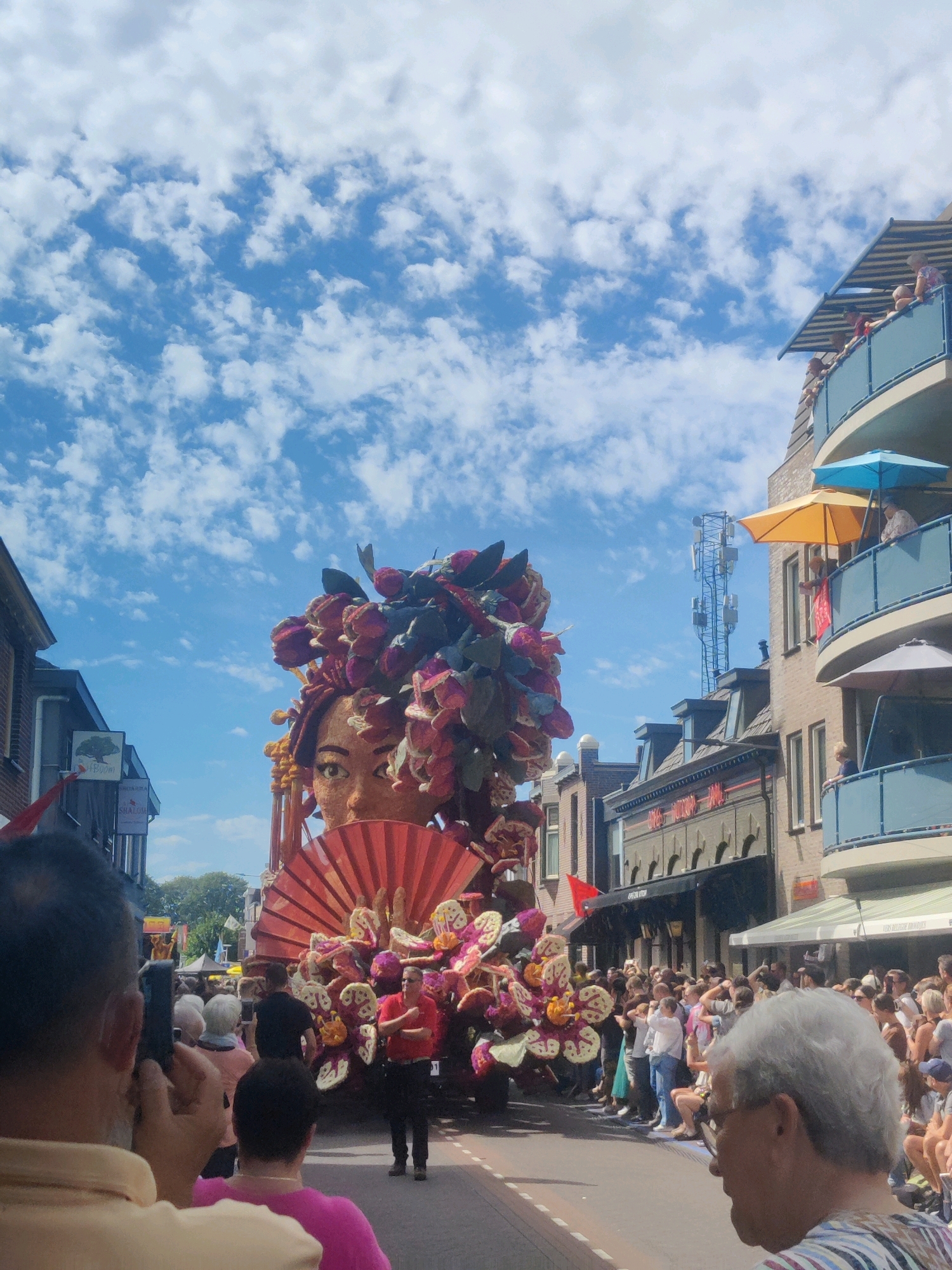 A large flower float depicting a woman's face with a fan is parading down a street in Zundert, Netherlands, during a flower parade. A crowd of spectators lines the street, some on balconies of the surrounding buildings, watching the parade. The sky is blue with scattered clouds. The float is adorned with numerous colorful flowers and foliage. A man in a red shirt and black pants, likely a parade organizer or staff, stands near the float. The buildings on either side of the street feature balconies and shops. The scene captures a festive community event with a focus on the elaborate floral artistry of the float.