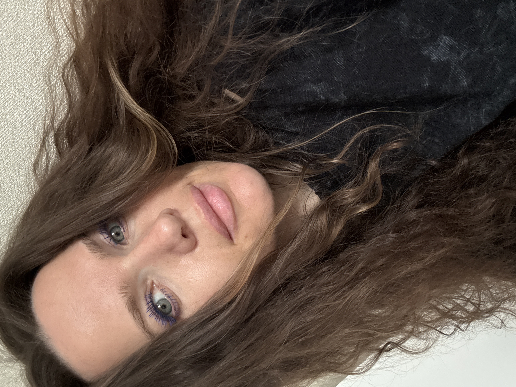 A close-up portrait shows a woman with light skin, gray-green eyes, and brown, wavy hair. The woman's face is slightly turned, with her head resting on a surface. She is wearing purple mascara. She appears to be lying down. The background consists of a textured white surface on one side and a black, textured garment on the other. The location is undetermined, and there is no indication that it is in Moscow, Russia.