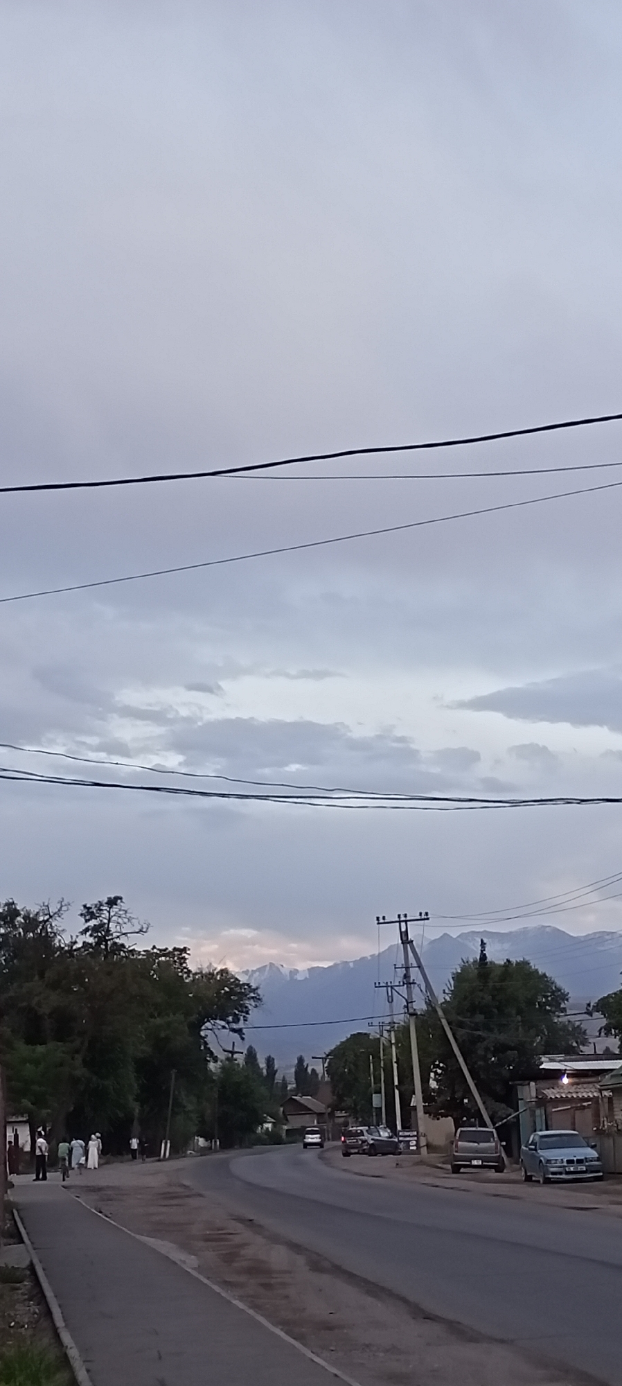 The image depicts a street scene in Sokuluk, Kyrgyzstan, under an overcast sky with prominent power lines stretching across the frame. A paved road curves through the scene, with a sidewalk on the left. On the sidewalk, several people are walking, including a group of individuals in what appears to be traditional attire. Trees line the left side of the road, and on the right, there are buildings and parked vehicles. In the distance, a range of mountains with snow-capped peaks is visible. A utility pole with wires stands near the right edge of the road, and other poles are scattered throughout the scene. Several cars are parked along the right side of the road and on the street. The overall atmosphere suggests a quiet, possibly late afternoon or early evening in a semi-urban or suburban area.