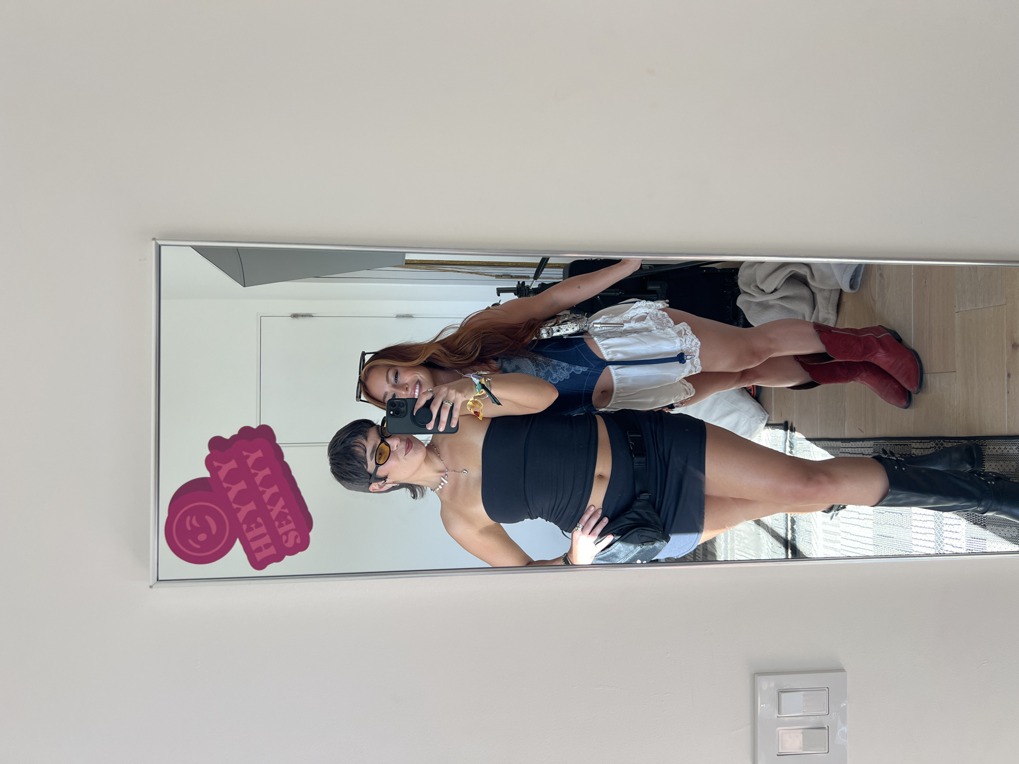 Two women pose for a selfie in front of a large rectangular mirror. The mirror is mounted on a white wall, beneath a light switch. The woman in the foreground wears a black tube top, a black mini skirt, and black knee-high boots. She also has a black fanny pack on her waist and yellow sunglasses. The woman in the background has long red hair and wears a blue lace-trimmed top, a white skirt, and red cowboy boots. A pink sticker with the words "HEYYY SEXYYY" is affixed to the upper left corner of the mirror. The location of the photograph is in a residential area of Indio, United States of America.