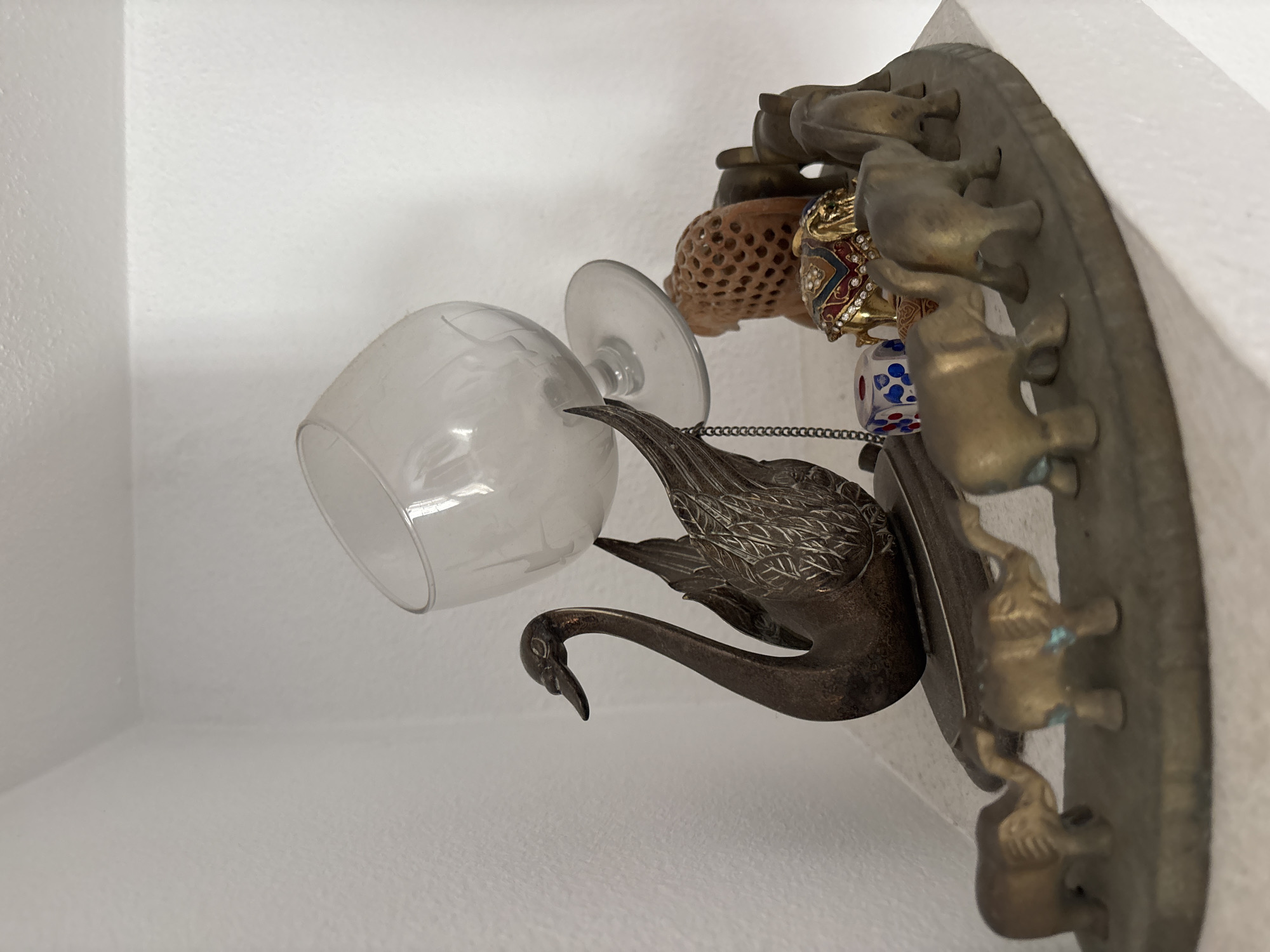 A small, tiered display shelf, likely made of concrete, holds a collection of decorative objects against a white textured wall. On the top tier, a clear glass brandy snifter is precariously balanced at an angle. To its left, a dark, metallic sculpture of a swan with intricate feather details is positioned. A thin, dark metal chain hangs from the swan's wing. To the right of the swan, a cluster of small, golden elephant figurines stands. One elephant is adorned with colorful rhinestones and metallic detailing. Behind the elephants, a light brown, porous decorative object, possibly carved wood or stone, is visible. Further to the right, three more golden elephant figurines are arranged. On the lower tier of the shelf, a line of smaller golden elephant figurines is depicted, each with a slightly different pose and coloring. One of these elephants has faint blue markings. The overall impression is of a curated display of various trinkets and decorative items. The location is not specified in the image.
