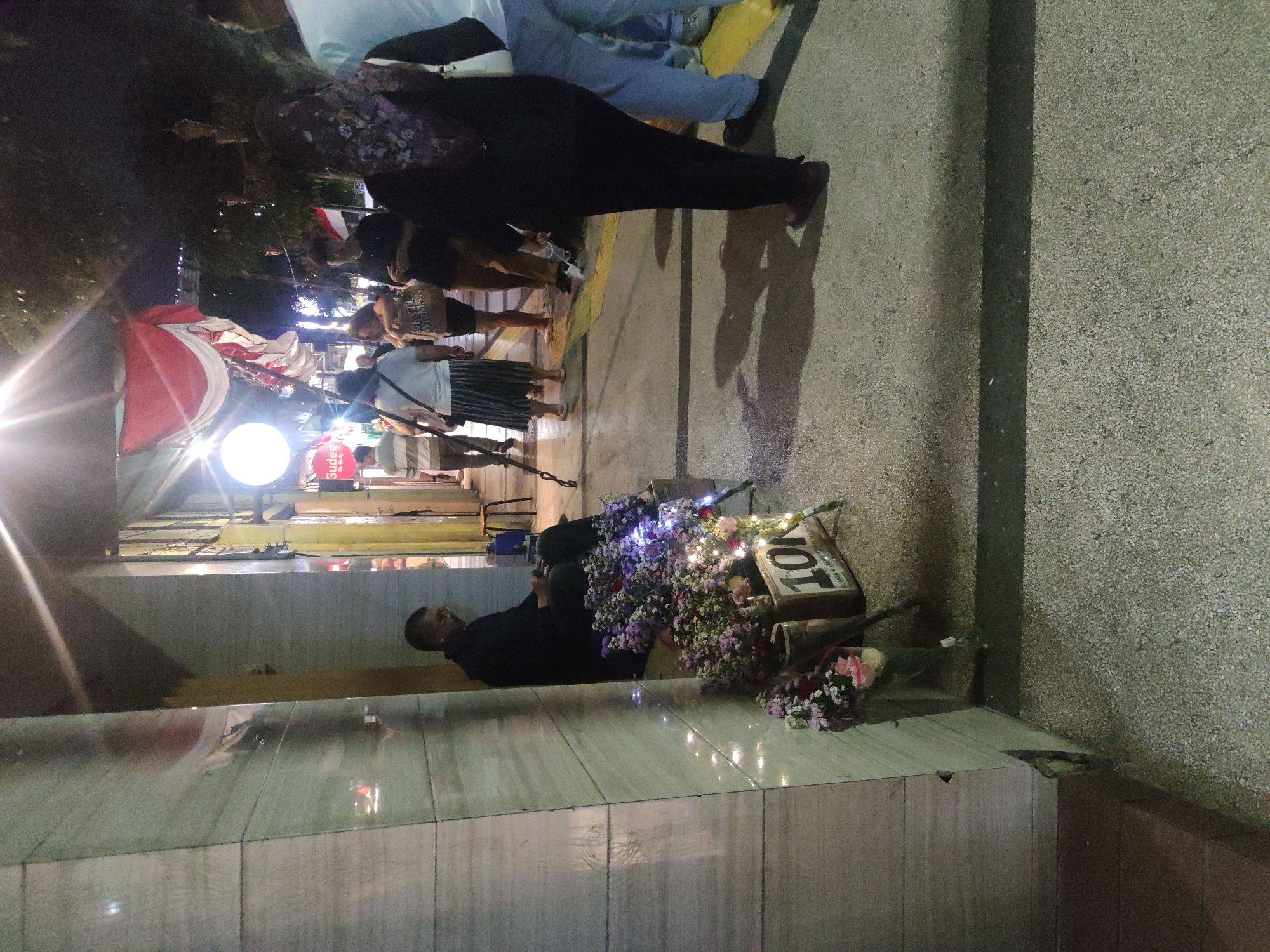 This is a nighttime, medium-angle, street-level photograph taken in the city of Malang, Indonesia. The scene depicts a man sitting on the ground, leaning against a building column, surrounded by numerous flower arrangements displayed in buckets and wooden crates. One wooden crate prominently displays the price "10K". The man is wearing dark clothing and glasses and appears to be looking down at his hands. String lights are draped around the building's base, adding a festive ambiance. In the background, several people are walking along a sidewalk, some with umbrellas. The buildings on the street feature decorative banners, including a red and white Indonesian flag. The ground is paved with gravel, and shadows are cast across it by the overhead lighting and the figures in the scene. The overall impression is one of a lively evening marketplace or street.