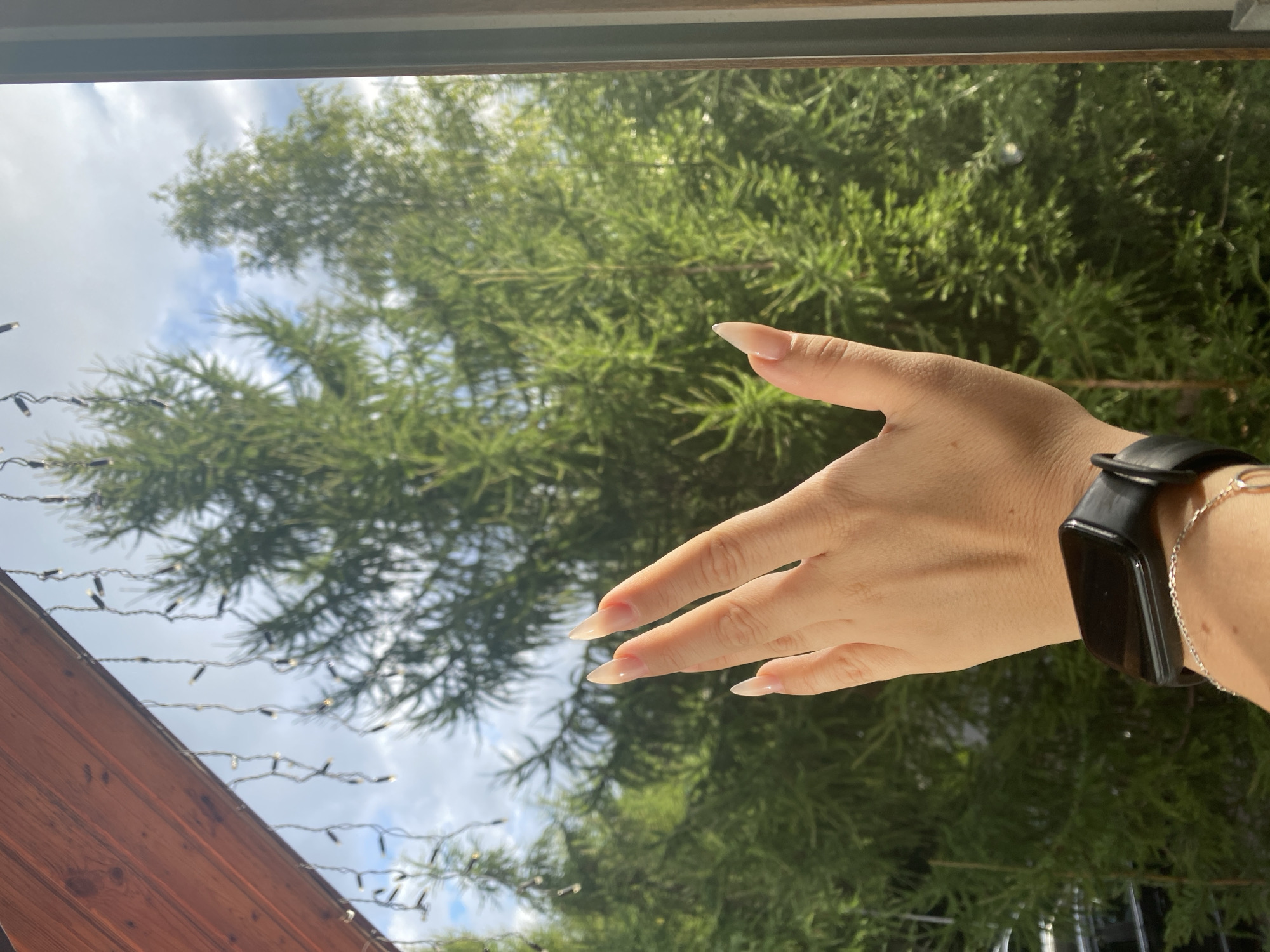 A close-up, medium shot captures a person's left hand and wrist against a backdrop of green trees and a cloudy sky. The hand, with long, almond-shaped, light-colored nails, is raised, fingers slightly splayed. The wrist is adorned with a black smartwatch and a delicate silver bracelet. The scene appears to be set on a balcony or porch, as indicated by a wooden beam and string lights visible in the upper left corner. The trees suggest a natural, possibly forested, environment. The image does not provide specific details to confirm its location in Krasnoflotskoe, Russia.