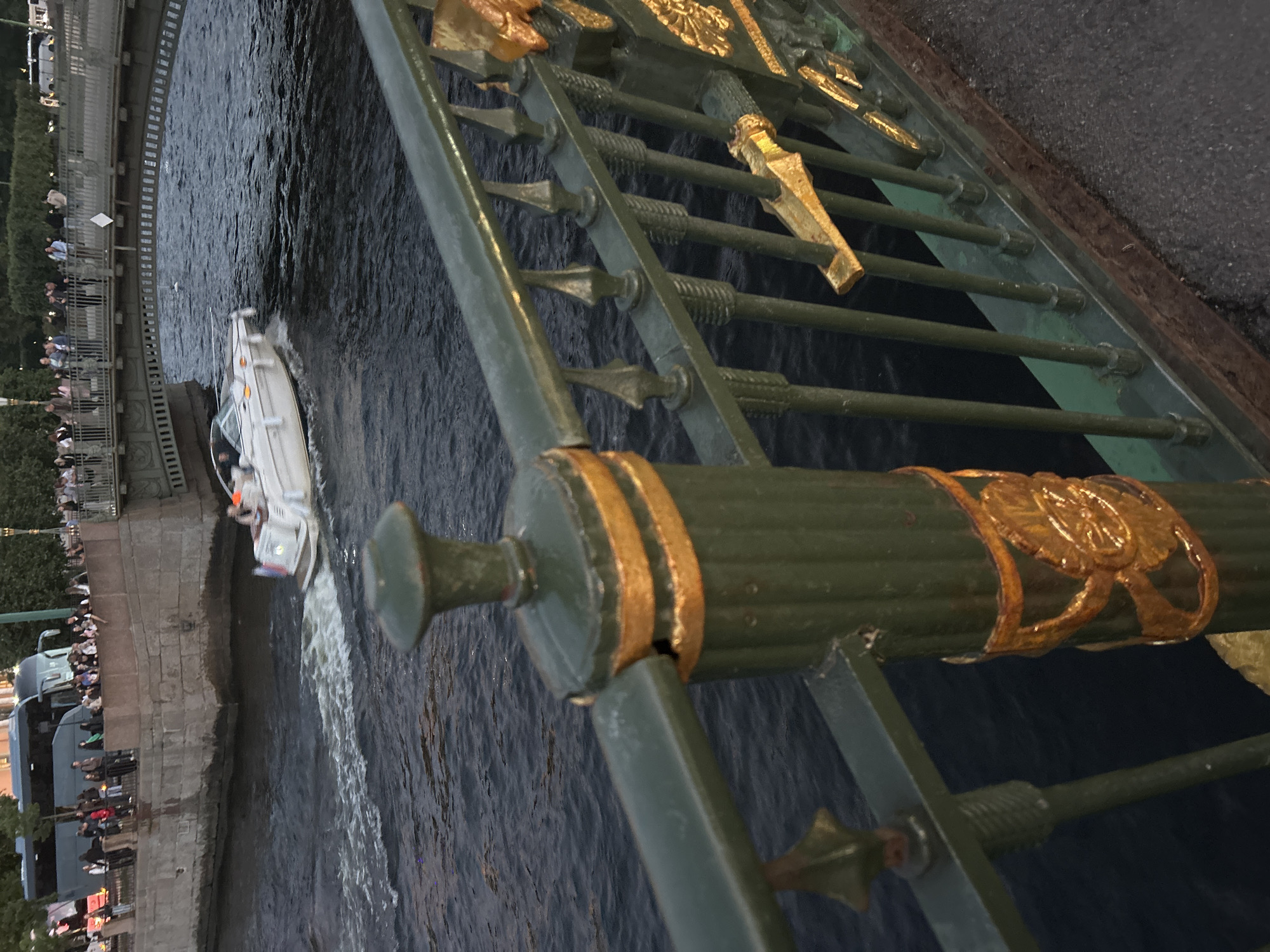 The image depicts a scenic view of a river and a bridge in an urban setting. In the foreground, a green and gold ornate metal railing runs across the bottom of the frame, suggesting the viewpoint is from a bridge or elevated walkway. The railing has decorative elements and a fluted column with a gold band.
In the middle ground, a white motorboat with a few occupants is speeding across the dark, rippling water, leaving a wake. The boat has a flag on its stern. Behind the boat, a stone embankment is visible, lined with people observing the scene. A large tour bus is parked on the embankment.
The background features a bridge with an arched structure, spanning the river. The bridge is also lined with people. Beyond the bridge, trees and buildings can be seen, indicating a developed urban environment. The overall lighting suggests it is either early morning or late evening, with diffused light.