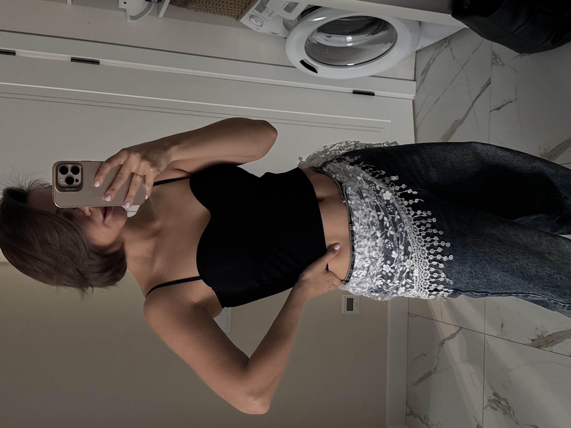 An individual is captured in a mirror selfie, posing indoors. The person, likely female, has short brown hair and is wearing a black strappy tank top or camisole paired with blue denim pants. The pants feature an elaborate white lace or embroidered overlay around the waist and upper thigh area, adding a decorative element to the outfit. One hand holds a light-colored smartphone (an iPhone with multiple cameras visible) to take the photo, revealing white nail polish and a ring on a finger. The other hand rests gently on her hip/waist.
The setting appears to be a modern interior space, possibly a utility room or bathroom, indicated by the presence of a white front-loading washing machine in the background. Above the washing machine, part of a woven basket is visible. The floor is covered with large white tiles featuring distinctive grey veining, resembling marble. White doors or cabinetry are also part of the background. A small white rectangular object, possibly a thermostat or light switch, is mounted on a wall. The lighting appears to be even, suggesting indoor illumination. The words "CAMERA" are faintly visible around the phone's camera lenses, and unreadable text is present on the phone case.