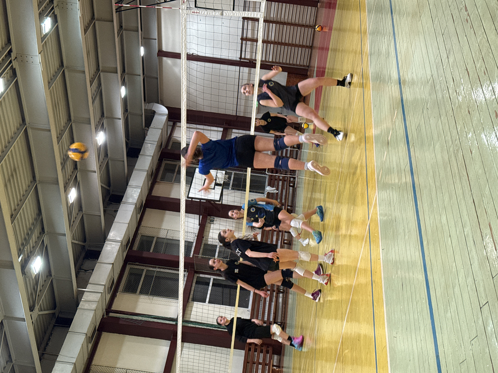 An indoor gymnasium in Sofia, Bulgaria, hosts a women's volleyball game or practice. Seven female athletes, clad in athletic shorts, t-shirts, and knee pads, are present on a polished wooden court featuring light green and yellow sections demarcated by blue lines. In the central foreground, one player in a blue t-shirt and black shorts is airborne, having executed a jump near the white volleyball net. A yellow and blue volleyball is suspended mid-air above her, exhibiting motion blur. Another player in a black sleeveless top and black shorts stands in a ready position in the foreground right. Additional players in black attire are positioned across the court, observing the play. The gymnasium walls are light-colored with prominent dark reddish-brown structural pillars and multiple large windows secured with metal grates. Tiered wooden benches line the left side of the court. The ceiling features an exposed industrial design with corrugated metal panels, structural beams, and multiple rectangular LED light fixtures. Horizontal metal ventilation ducts run along the upper walls. Two additional volleyballs are visible on the court floor in the background. The facility's appearance is consistent with an institutional or public sports hall.