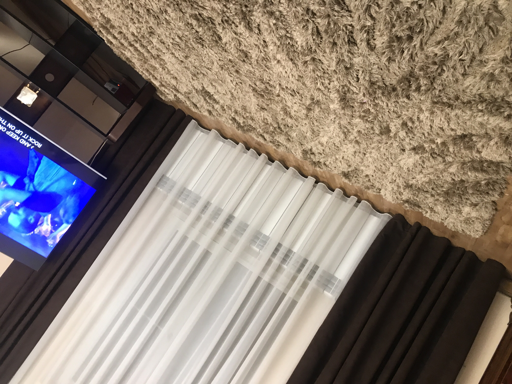 The image shows a room interior. The main elements are a television with a bright blue screen, brown blackout curtains, sheer white curtains, a beige shag rug, and a dark-colored media cabinet. The curtains cover a window. "ROCK IT UP ON THE" is visible on the TV screen. The floor is made of wood. The image is taken at an oblique angle. There is no indication of the location being Yerevan, Armenia.