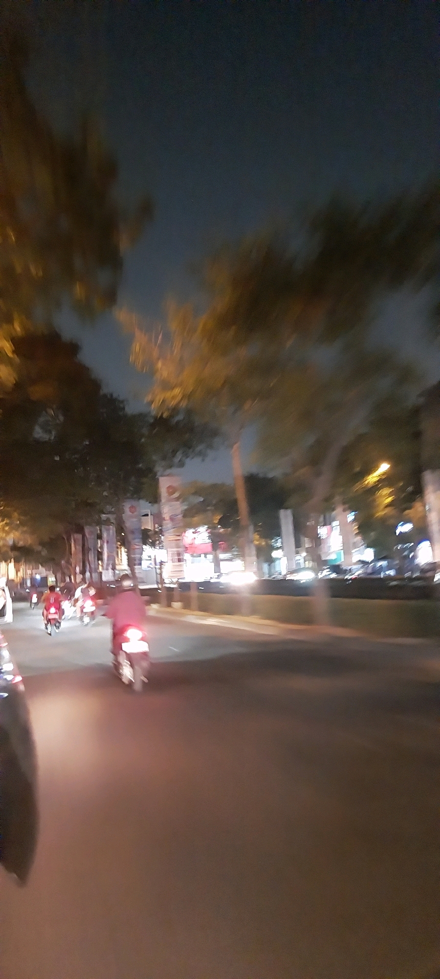 The image captures a blurred nighttime street scene in Kelapa Dua, Indonesia. Several motorcycles are visible, with riders in motion. One motorcycle in the foreground has its taillight illuminated. Trees line the street, and in the background, there are blurred lights from buildings and streetlights. The sky is dark, indicating it is nighttime. The perspective suggests the image was taken from a moving vehicle.