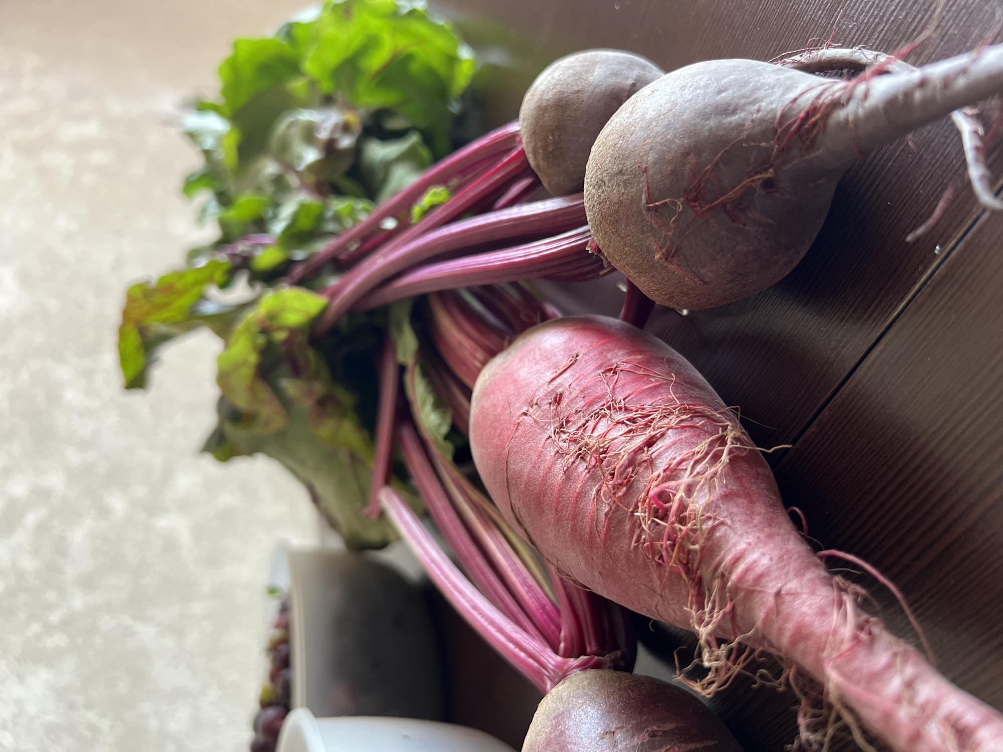 A bunch of raw beets with green tops lies on a dark wooden surface. The beets are a deep red and purple color, with thin roots trailing from them. In the background, there is a white bowl filled with what appear to be olives. The environment is indoors, with a light-colored textured wall behind the subjects. The image is a close-up shot, focusing on the vegetables. There are no people or animals present in the image. The location is not specified in the image, and therefore cannot be identified as Tolyatti, Russia.