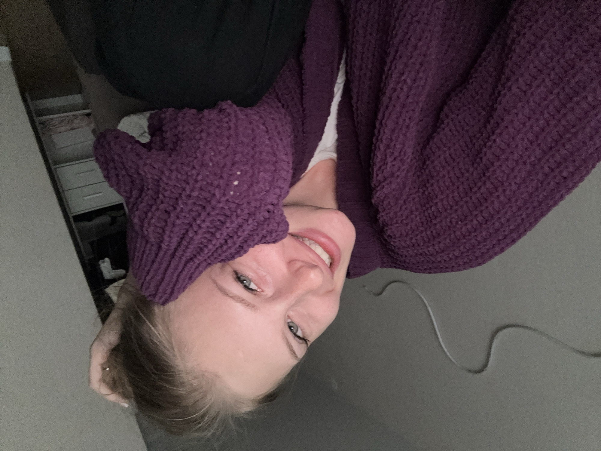 A young woman is shown from the chest up, resting her head on her hand, with her gaze directed towards the camera. She has fair skin, light blue eyes, and blonde hair that is pulled back. She is wearing a purple knitted cardigan over a white top. Her right arm is bent, and her hand is touching her head. In the background, a wall with a visible wire is present, as is a shelf with drawers and folded items. The image is a selfie taken indoors.