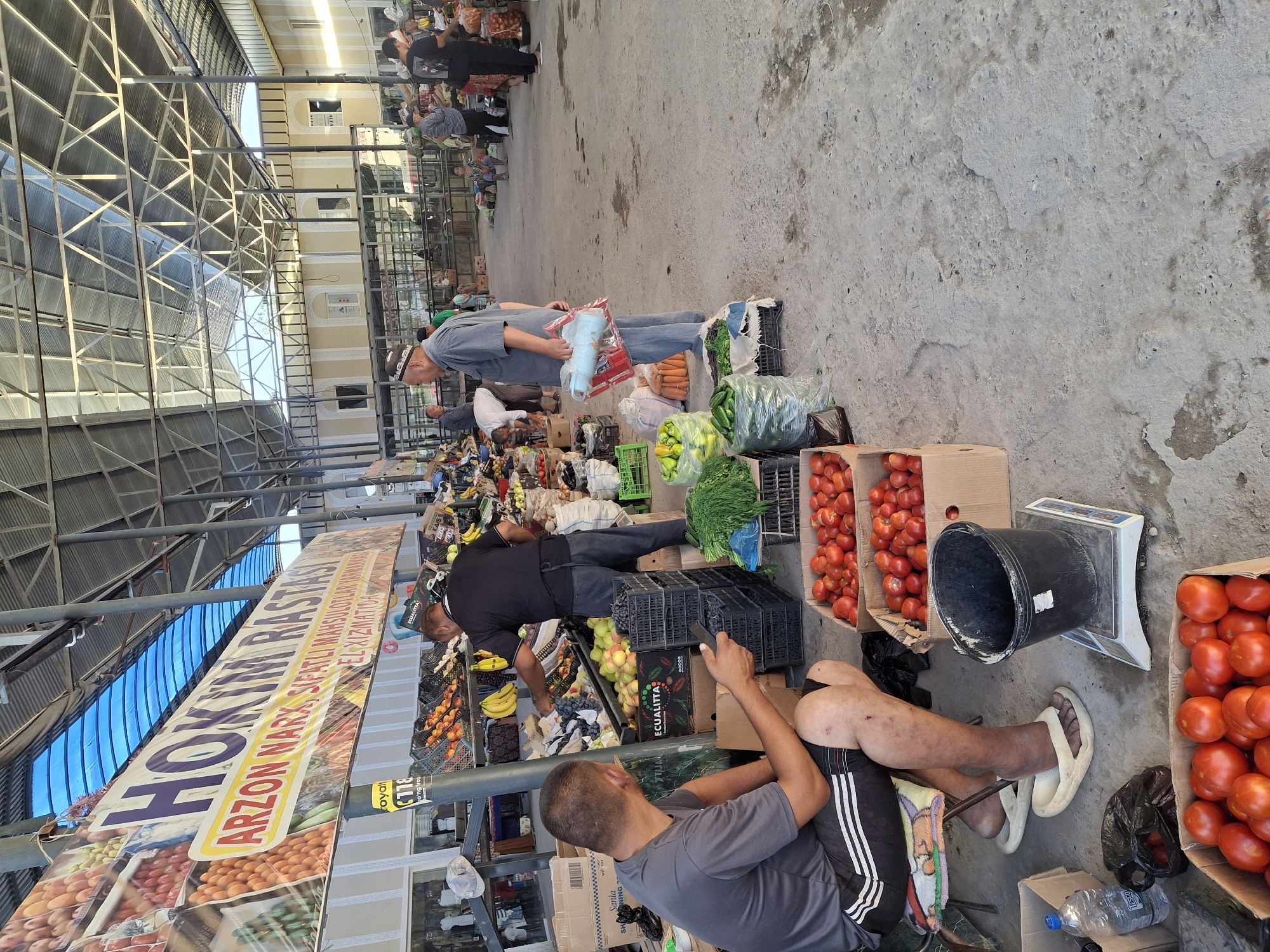 The image shows an outdoor market scene in Ko`kdala, Uzbekistan. A man in a grey t-shirt and black shorts is sitting on a stool, holding a mobile phone and looking down. In front of him are several boxes filled with red tomatoes. To his right, a black bucket sits on a scale. Another man in a black t-shirt and grey pants is standing behind a stall filled with fruits and vegetables, appearing to be serving a customer or arranging produce. The market stalls are set up under a large, open-air roof structure with metal beams and a translucent blue canopy. Various fruits, vegetables, and other goods are displayed for sale. Several other people are visible in the background, some browsing, some working, and some standing and talking. The ground is paved with concrete. A banner with text in a foreign language hangs above the stalls.