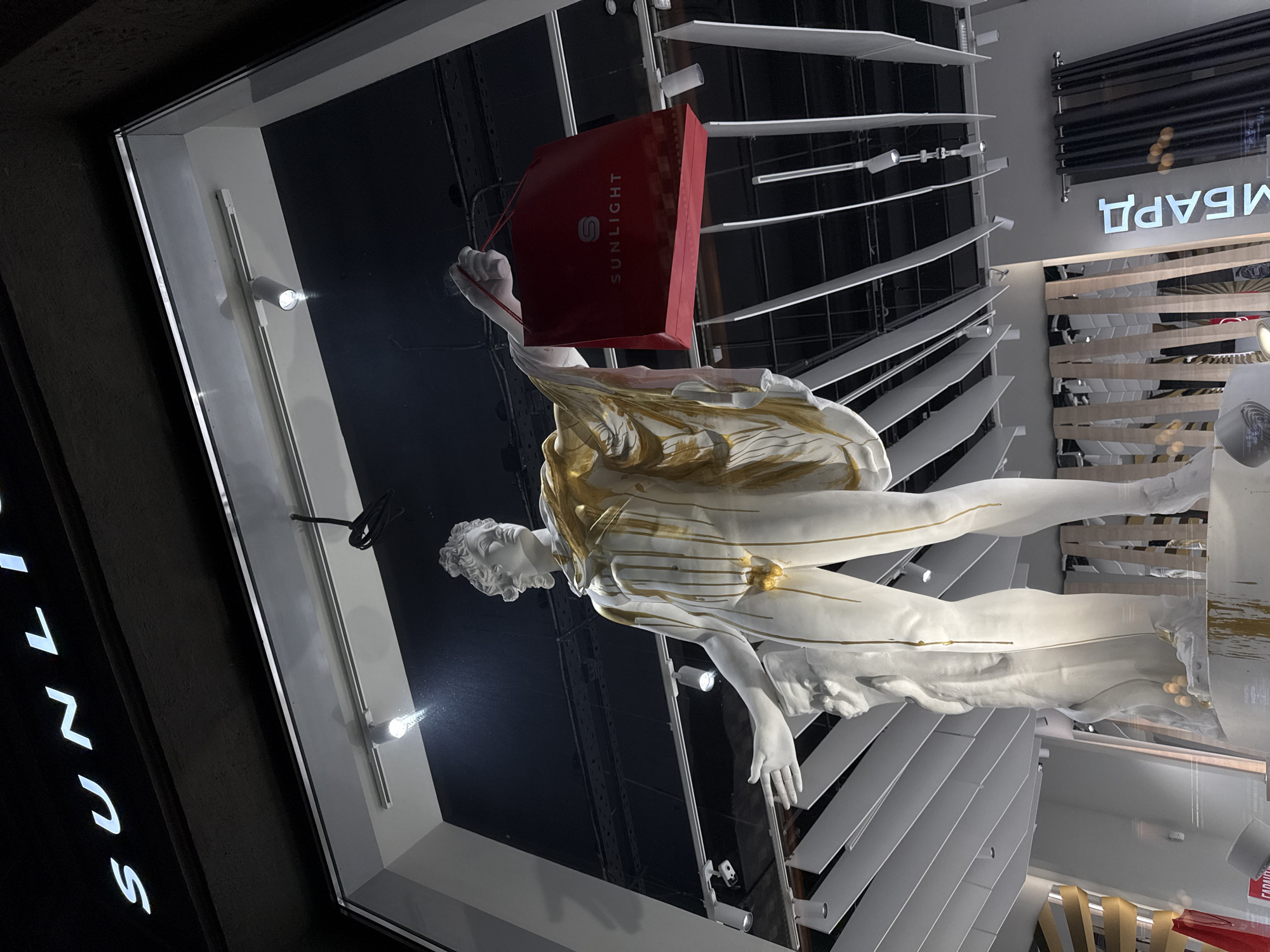 The image shows a storefront window display. A white and gold-accented statue of a male figure holds a red shopping bag with the word "SUNLIGHT" printed on it. Behind the statue are horizontal white slats resembling blinds. The store's name, "SUNLIGHT," is vertically spelled out in white letters on the dark exterior of the building. "BAPBA" is spelled out on the top right of the window. The location is Moscow, Russia.