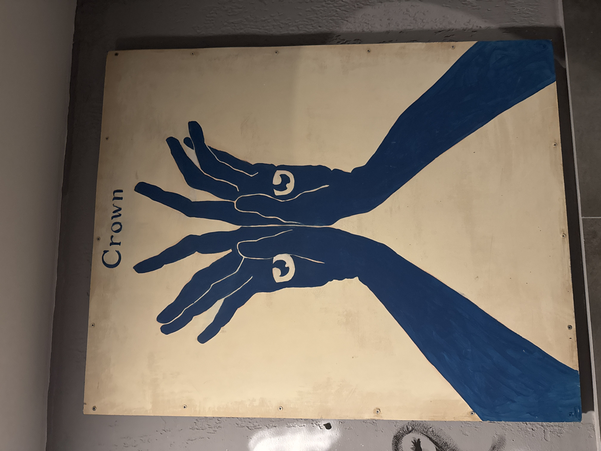 A rectangular art panel features a graphic depiction of two stylized hands in solid dark blue against a cream background. The hands are mirrored horizontally, with fingers spread and palms facing each other, angled slightly upwards. Each palm contains a distinctive white, ring-like shape, resembling an eye or a spiral. On the left side of the panel, oriented vertically, is the word "Crown" in dark blue capital letters.
The panel is affixed to a textured wall, likely concrete or painted grey, with visible screws in its corners and along its edges. The wall area below the sign shows indistinct markings or graffiti. The lighting suggests an indoor or sheltered outdoor setting. The artwork's style is bold and symbolic, potentially representing a gesture, a shadow puppet, or an abstract form of a crown created by the hands. This piece is located in Tbilisi, Georgia.