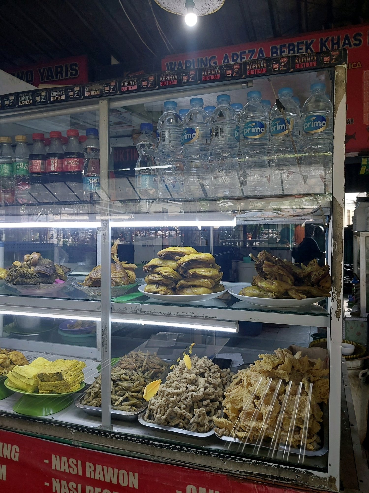 The image shows a food stall with a glass display cabinet filled with various food items and drinks. The stall appears to be located in Malang, Indonesia, as indicated by the red banner at the bottom which reads "- NASI RAWON - NASI D..." in Indonesian.
In the display cabinet, on the top shelf, there are numerous bottles of "Prima" brand bottled water. Below the water bottles, the middle shelf contains several dishes of cooked food. On the left, there are dishes of what appears to be tripe and offal. In the center, there are plates of cooked chicken. On the right, there are also plates of cooked chicken.
The bottom shelf of the display cabinet showcases more food items. On the left, there are slices of yellow food, possibly fried tofu or tempeh. Next to it are trays filled with small, fried fish or anchovies. To the right of the fish, there is a large tray filled with crispy, golden-brown fried food, possibly chicken or seafood.
Above the display cabinet, there are red signs with white text. One sign reads "YARIS", and another larger sign has text that appears to include "APREZA BEBEK KHAS" which translates to "Special Duck". There are also smaller repeated signs with "KOPIANYA BUKTIKANI!" which means "Try our coffee!"
The environment suggests an outdoor or semi-outdoor setting, typical for a street food stall in Indonesia. The lighting appears to be a mix of fluorescent lights within the display cabinet and overhead lighting. People are visible in the background through the glass, suggesting activity and customers. The overall scene depicts a bustling food stall ready to serve customers.